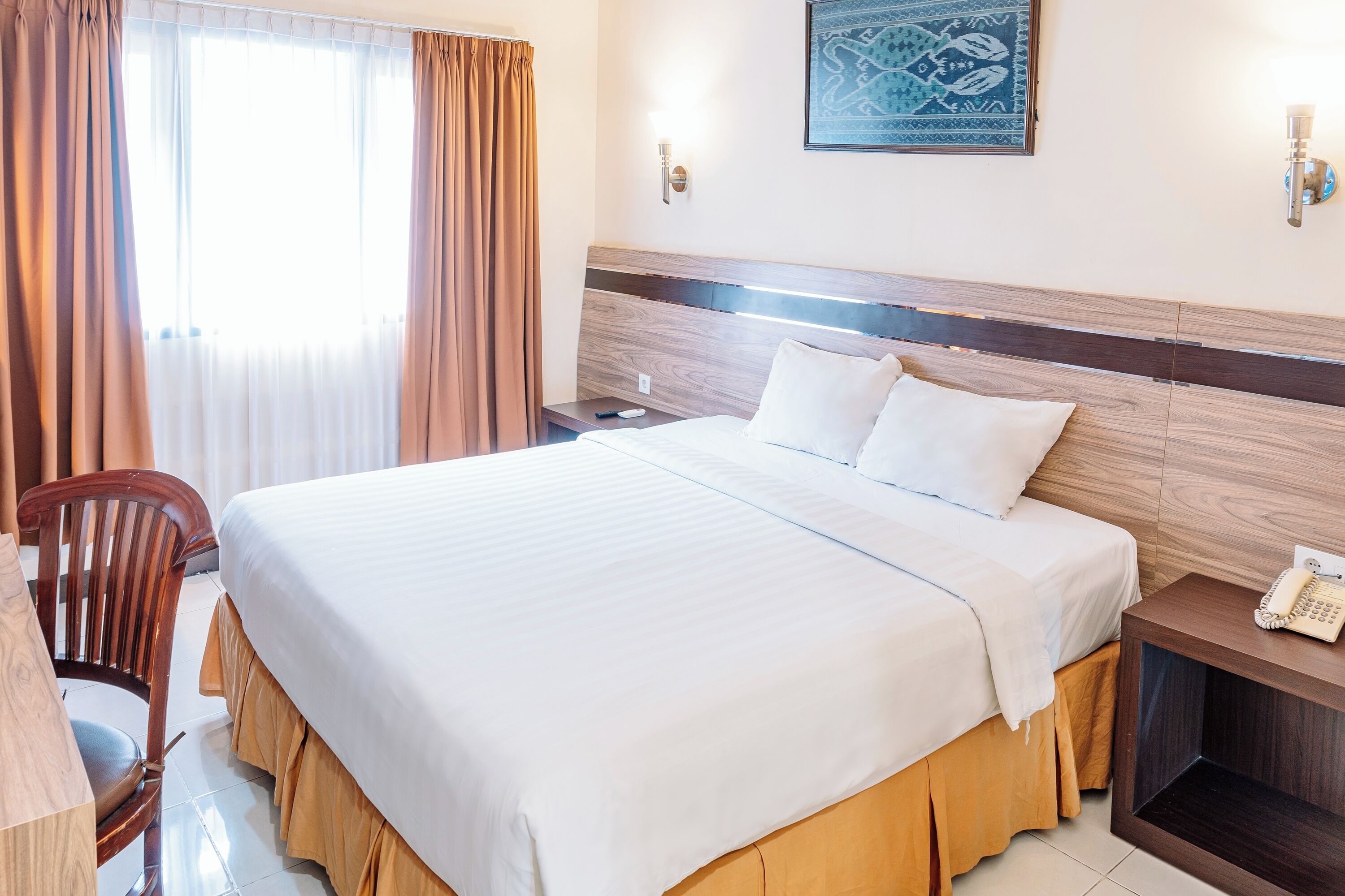 A hotel room with a bed, a chair, a bedside table with a phone, and a wall-mounted light fixture.