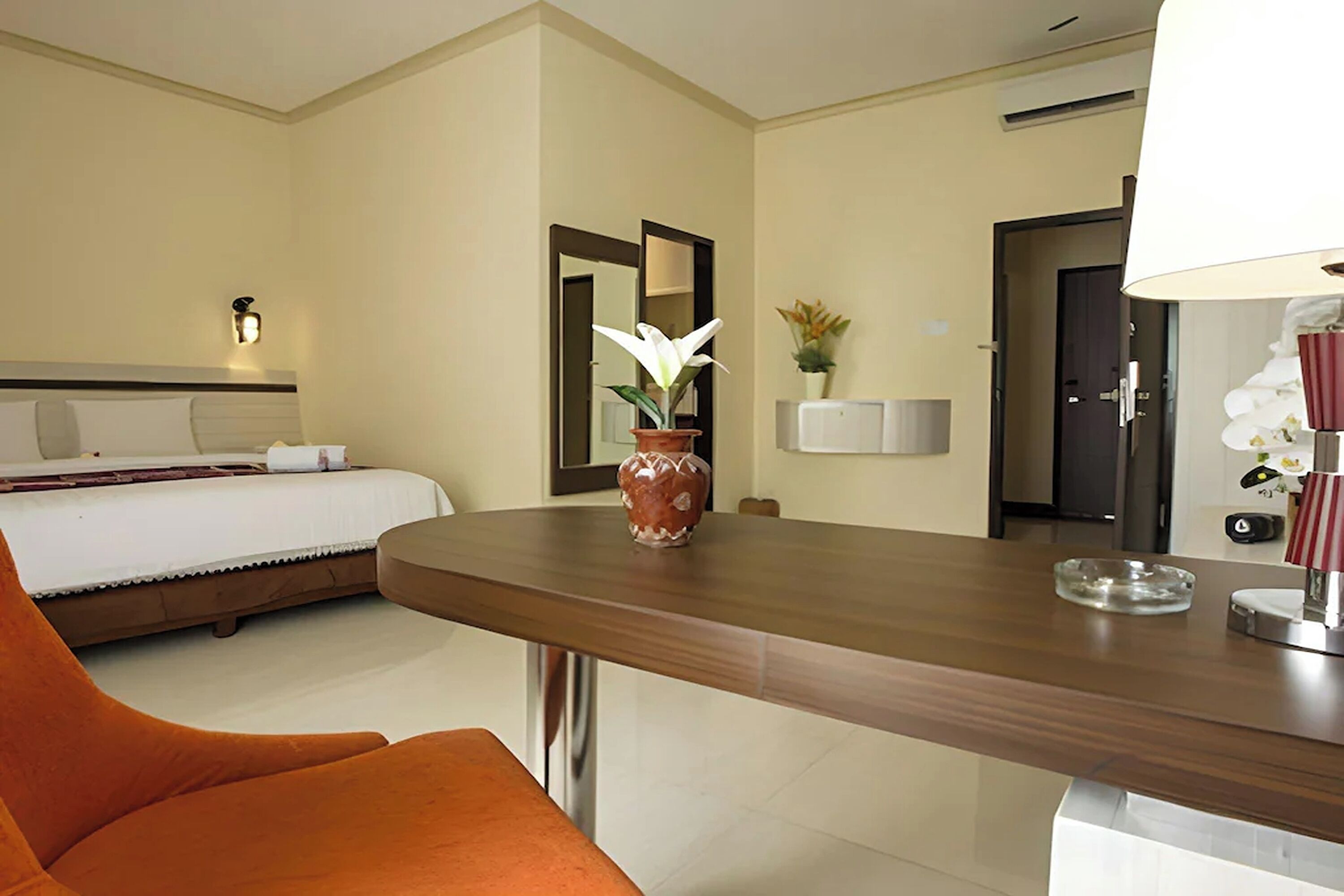 Habitación de hotel con una cama, un escritorio de madera, un florero con una flor, un espejo y una lámpara.