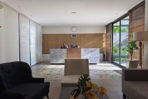 A reception area with a desk, chairs, a clock, and a view of greenery through large windows.
