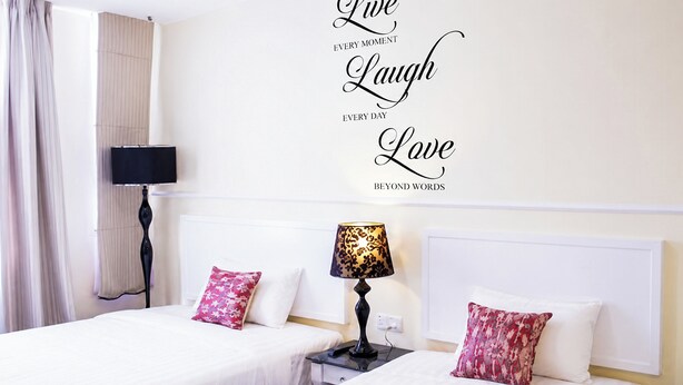 Habitación de hotel con cama, dos almohadas, mesita de noche, lámpara, televisor y una pared con texto inspirador.
