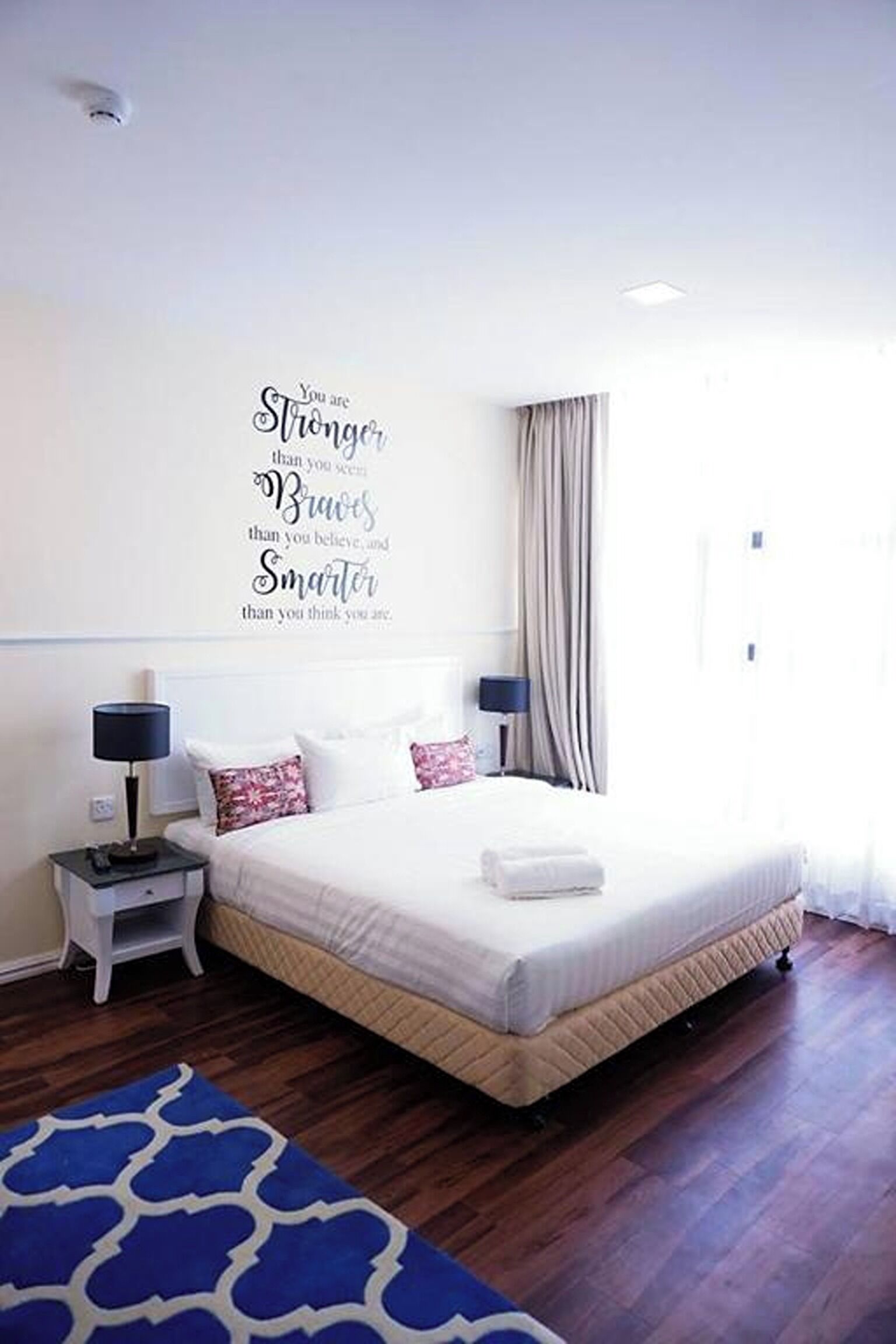 A bedroom with a bed, nightstands, a wall decor with motivational text, a window with curtains, and a blue rug.