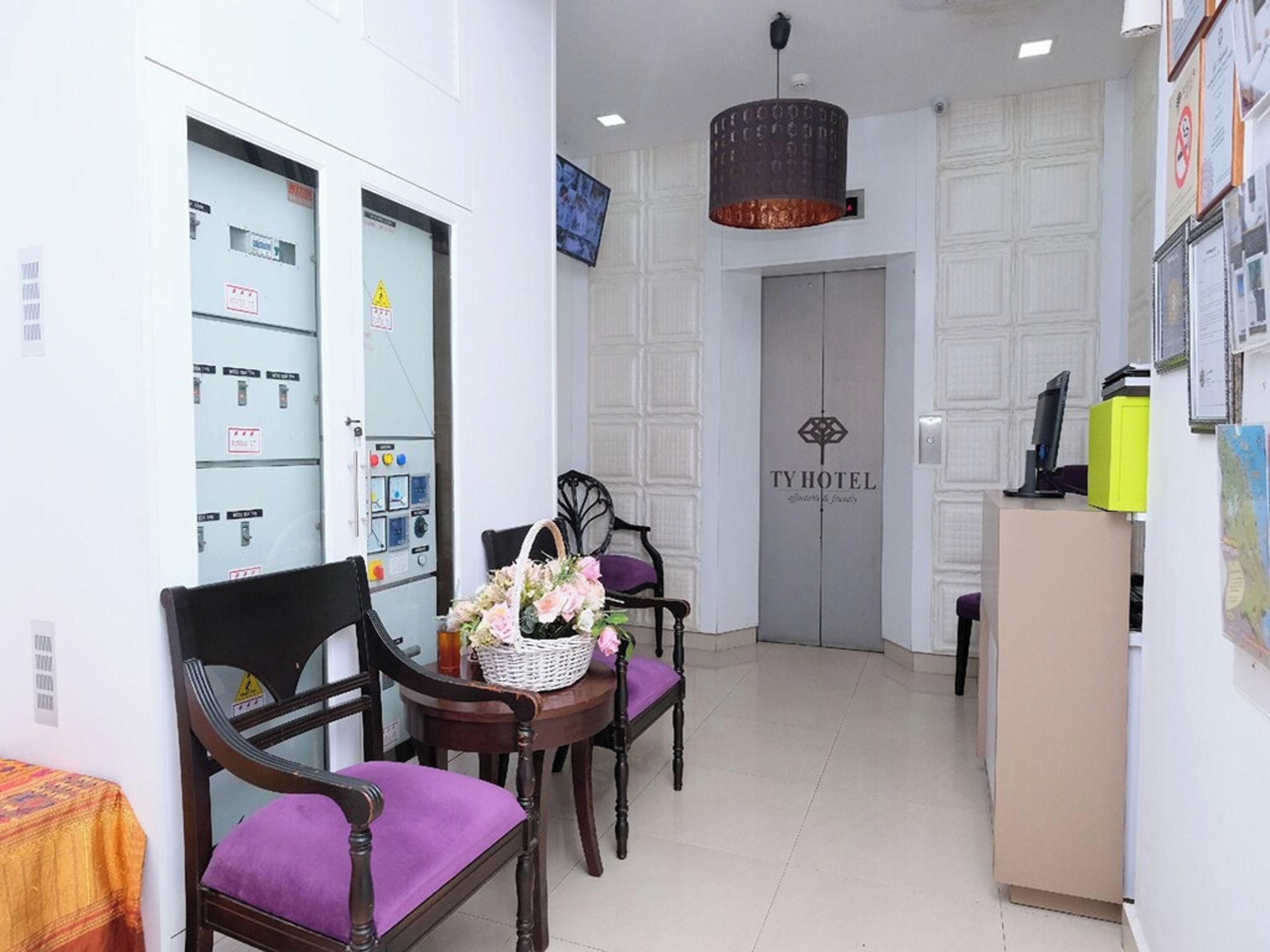 A hotel reception area with a reception desk, two chairs with purple cushions, a wall-mounted television, and a door with the hotel name.