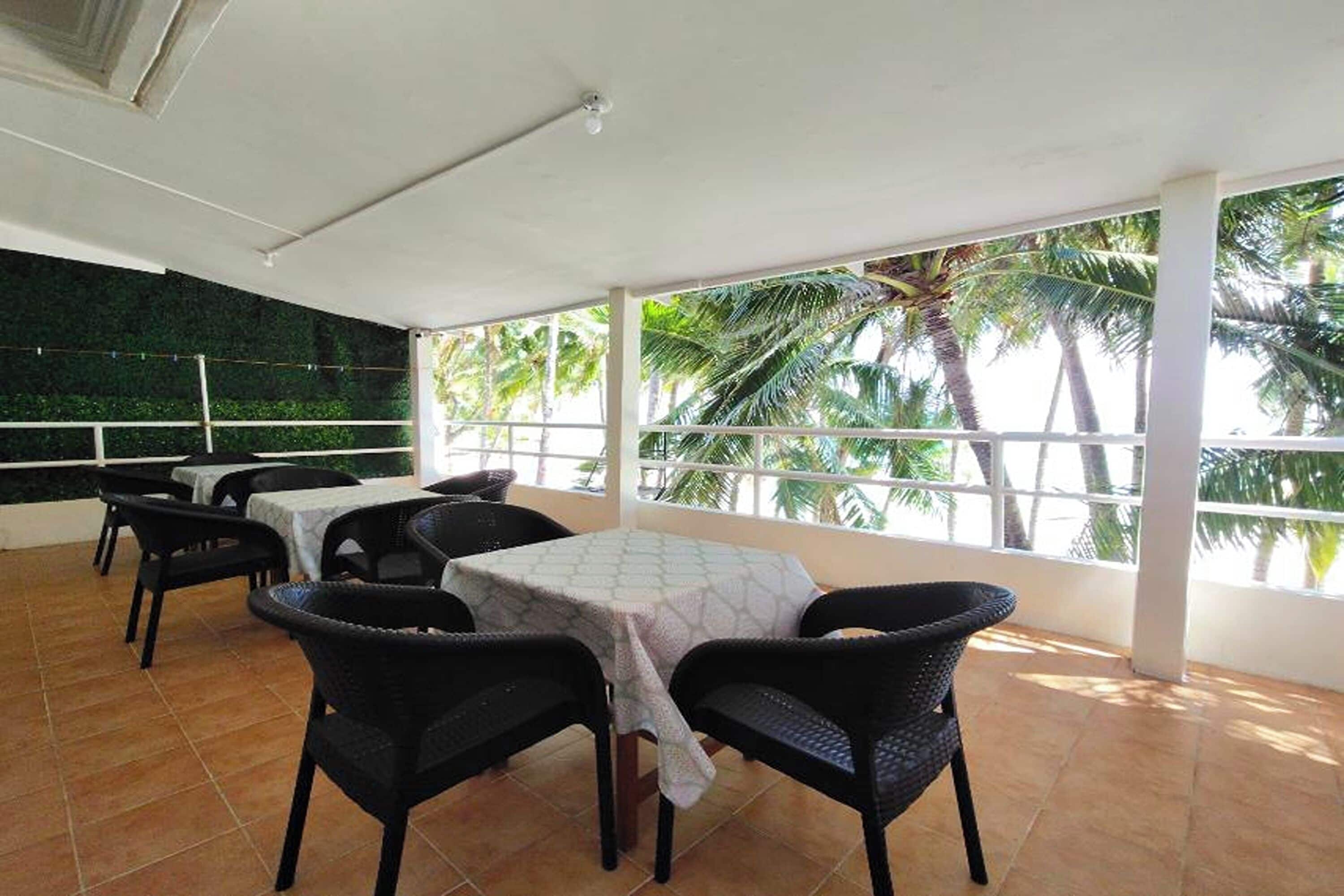 A covered outdoor dining area with tables and chairs, large windows, and a view of palm trees.