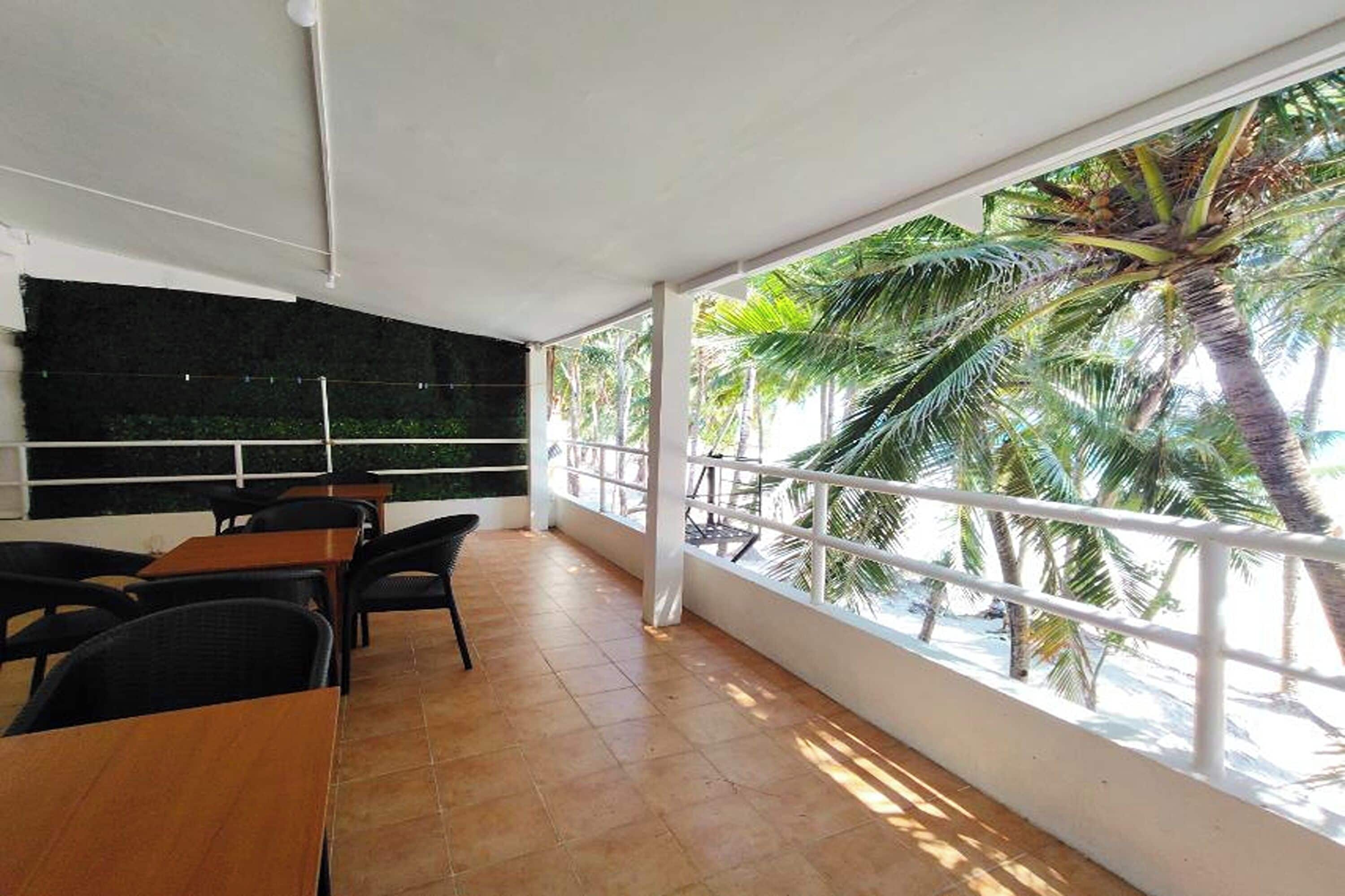 Un espacio al aire libre techado con mesas y sillas, ventanas amplias con vista a palmeras y piso de baldosas.