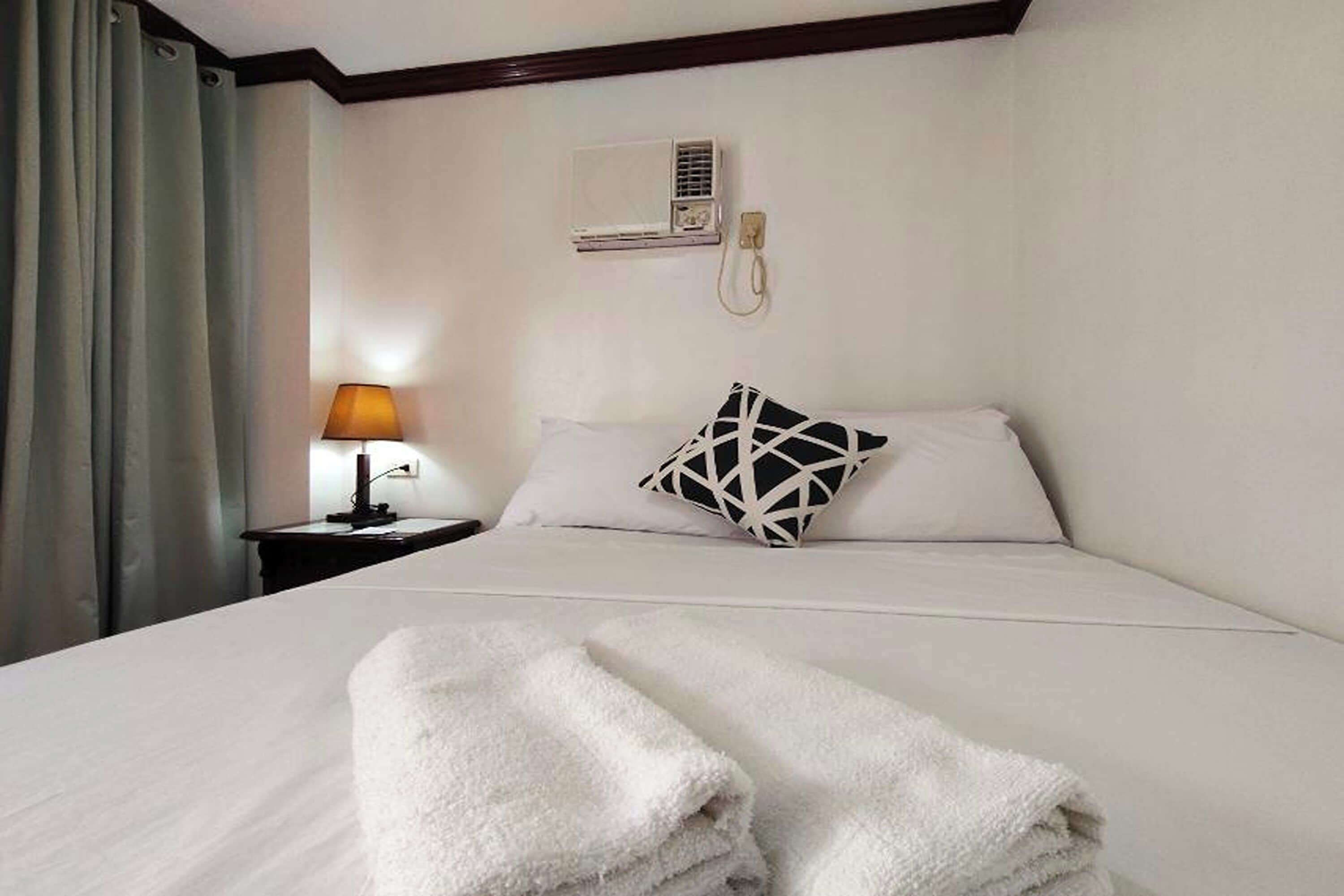 Habitación de hotel con cama, mesita de noche con lámpara, teléfono en la pared y ventana con cortinas.