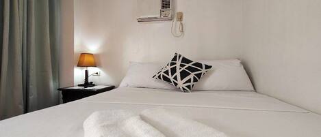 Habitación de hotel con cama, mesita de noche con lámpara, teléfono en la pared y ventana con cortinas.