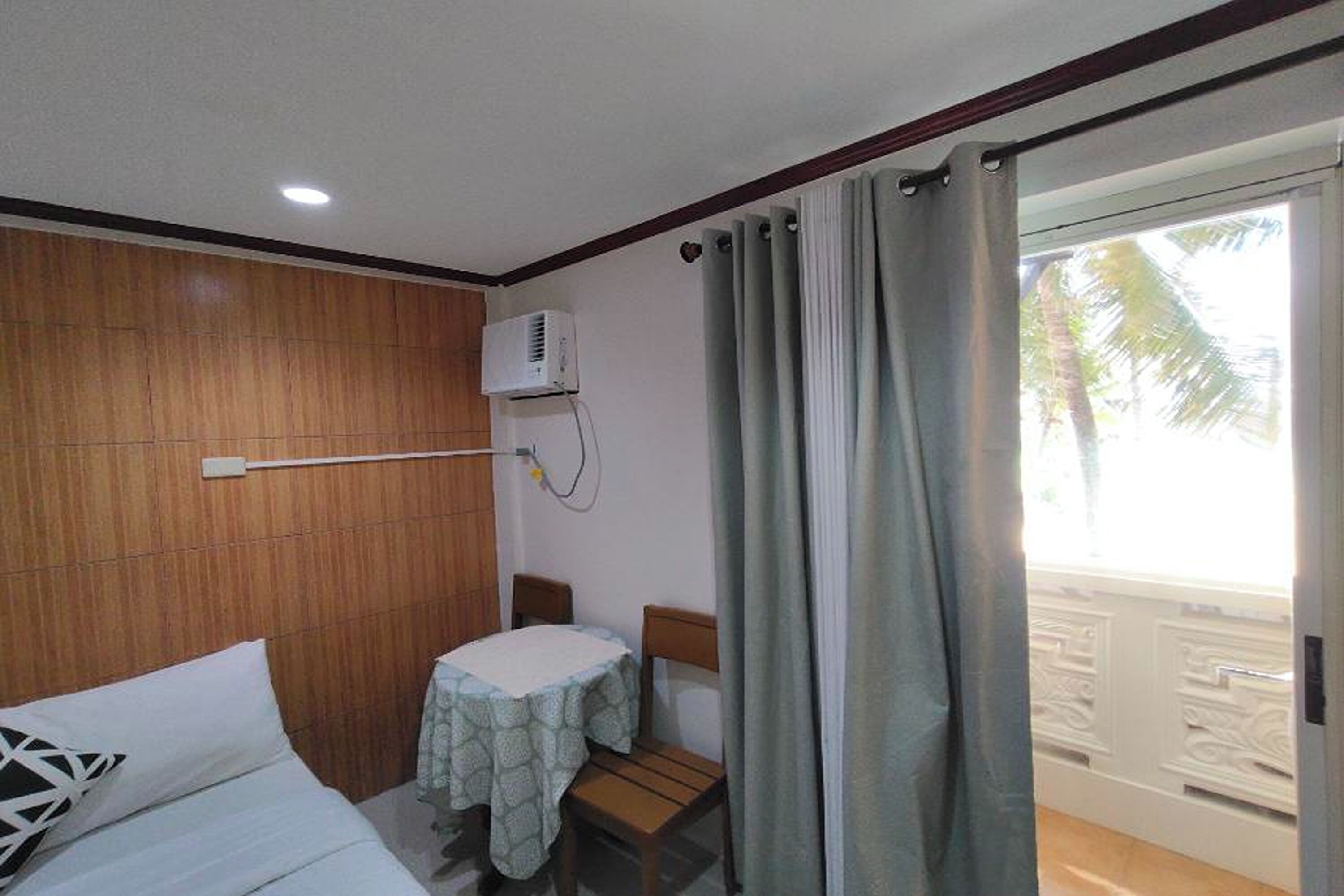 A hotel room with a bed, a table with a chair, an air conditioning unit, and a view of palm trees through the window.