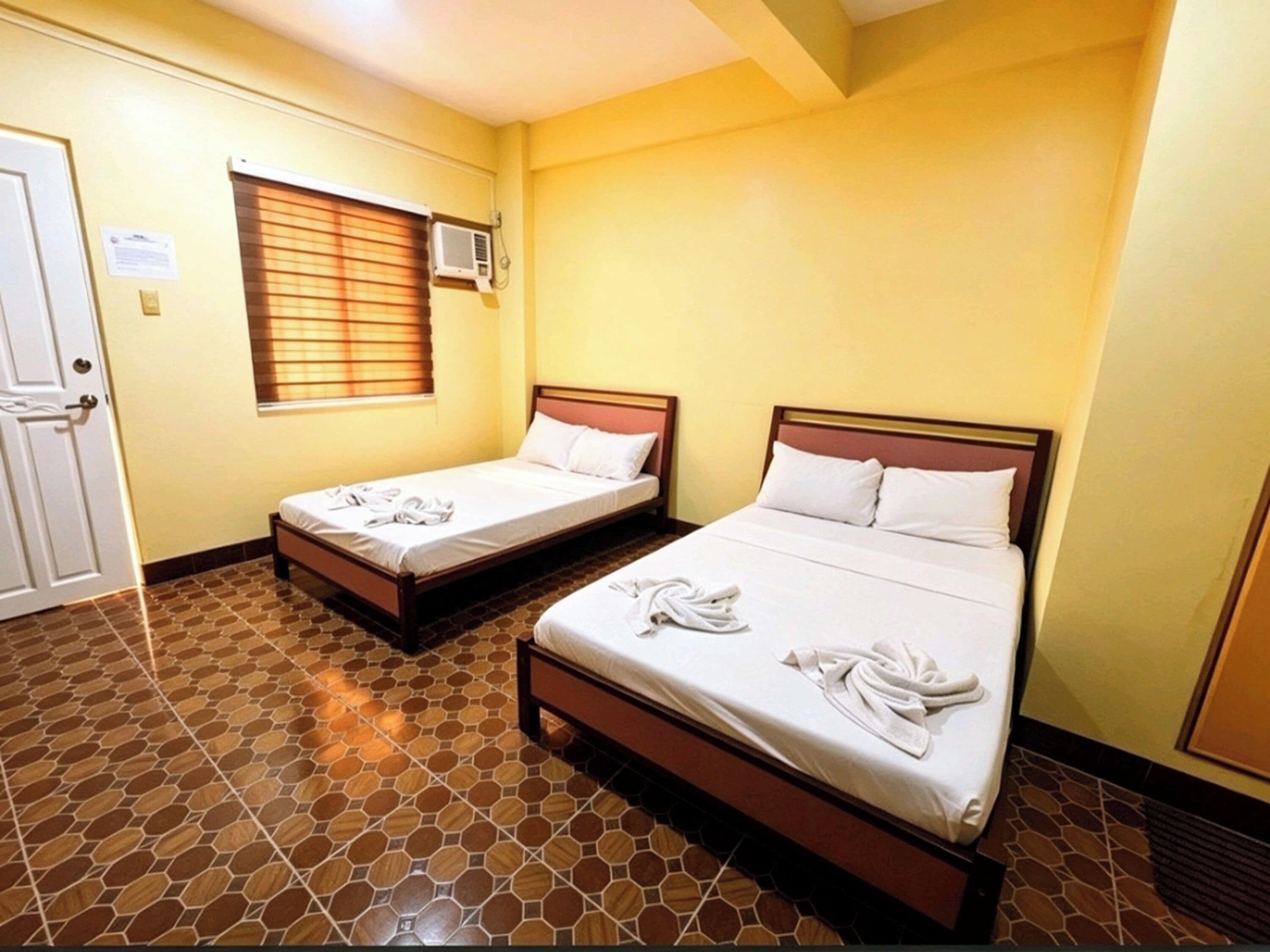 A hotel room with two single beds, a window with blinds, an air conditioning unit, and a tiled floor.