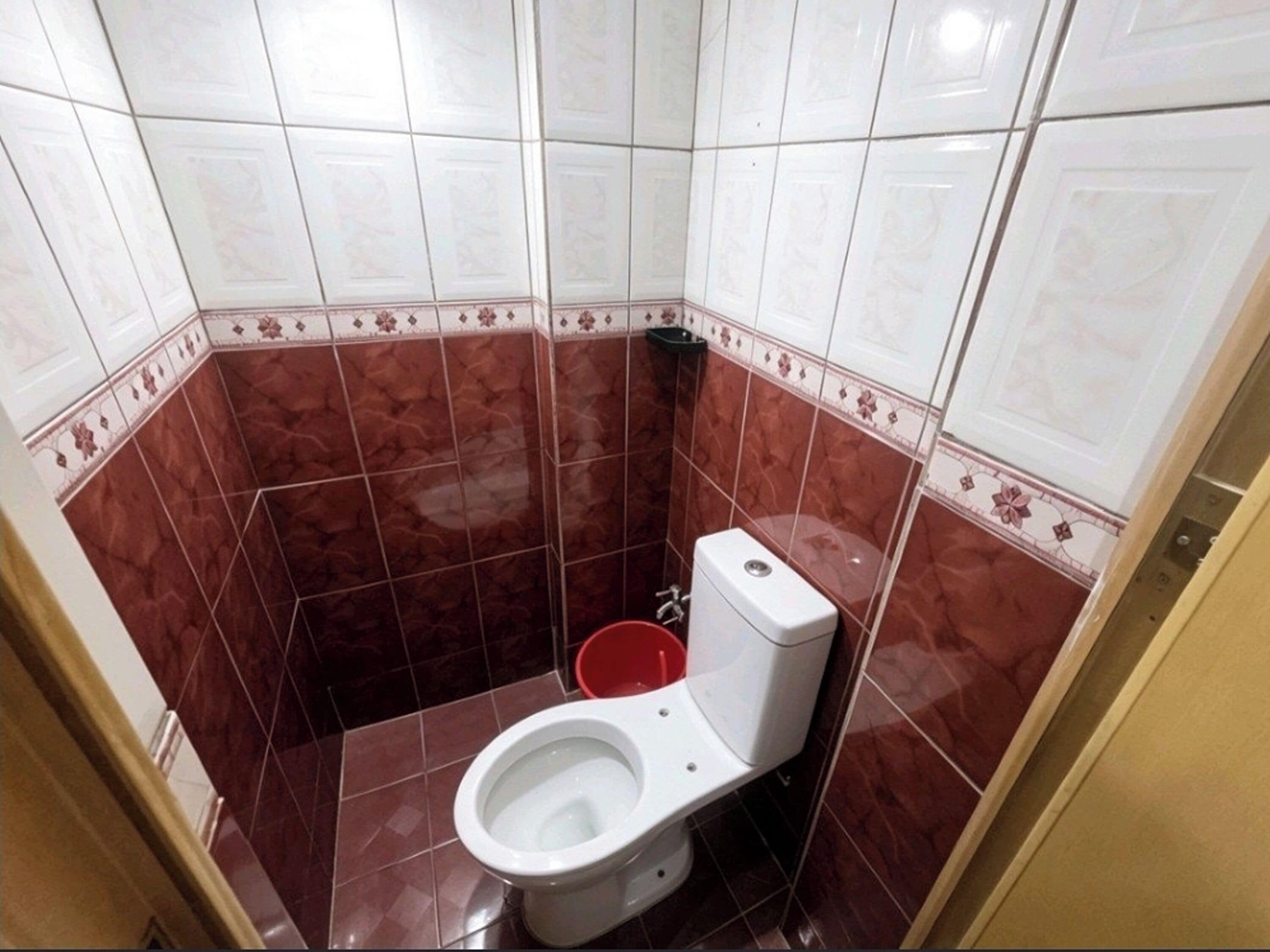 A small bathroom with a toilet, a red bucket, and a corner sink.
