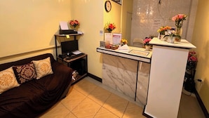 A hotel room with a bed, a desk with a computer, a clock, and a reception desk with flowers. - Nook Airport Guesthouse (Pasay)