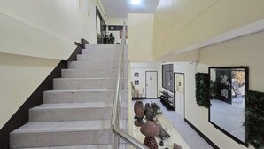 Una escalera con barandilla, una puerta con la etiqueta 'C' y un espejo que refleja una habitación con una planta.