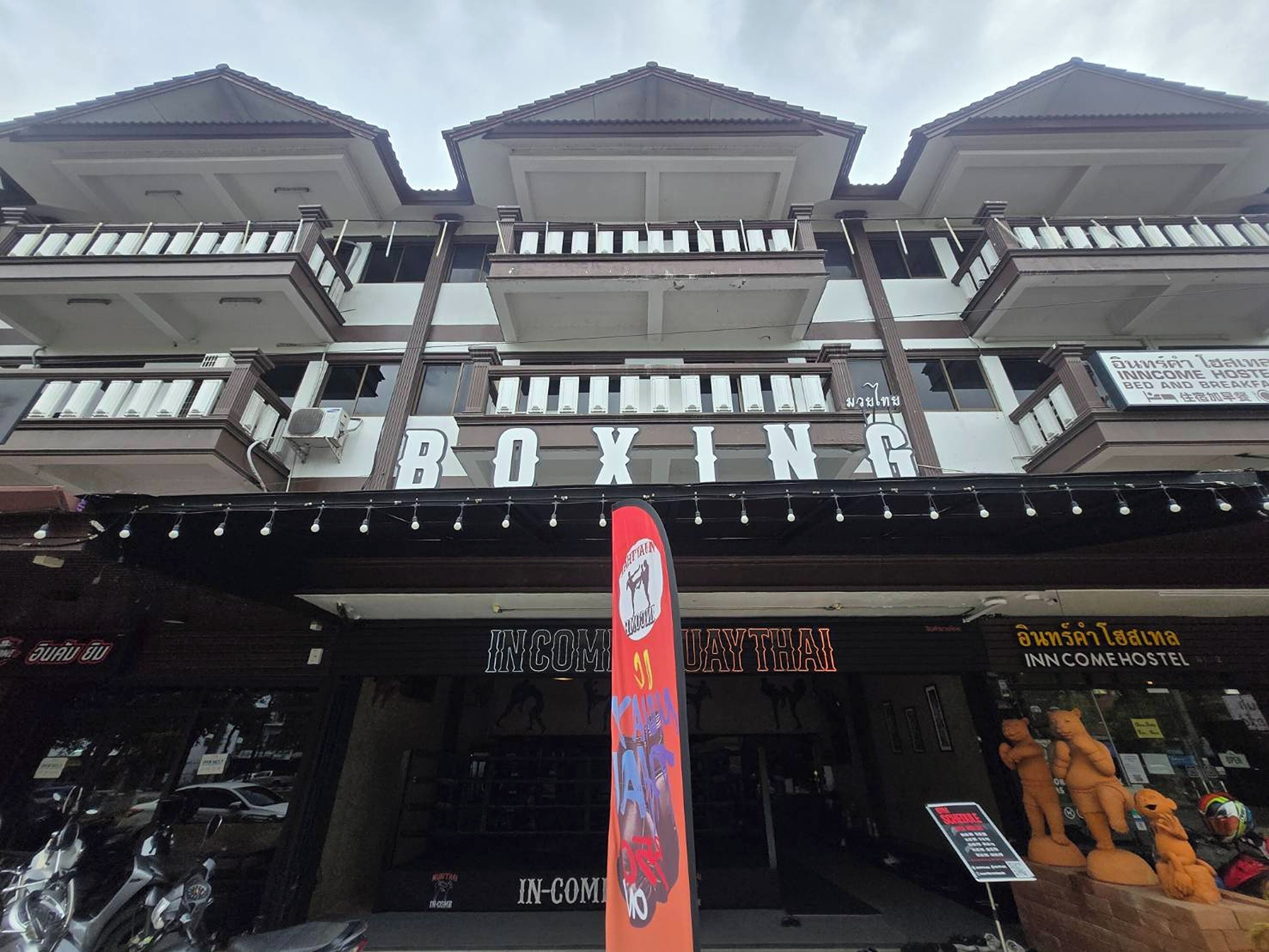 Um edifício com uma placa indicando 'BOXING INCOME HOSTEL' e 'DAY THAI', revelando um hostel com temática de boxe e um restaurante tailandês.