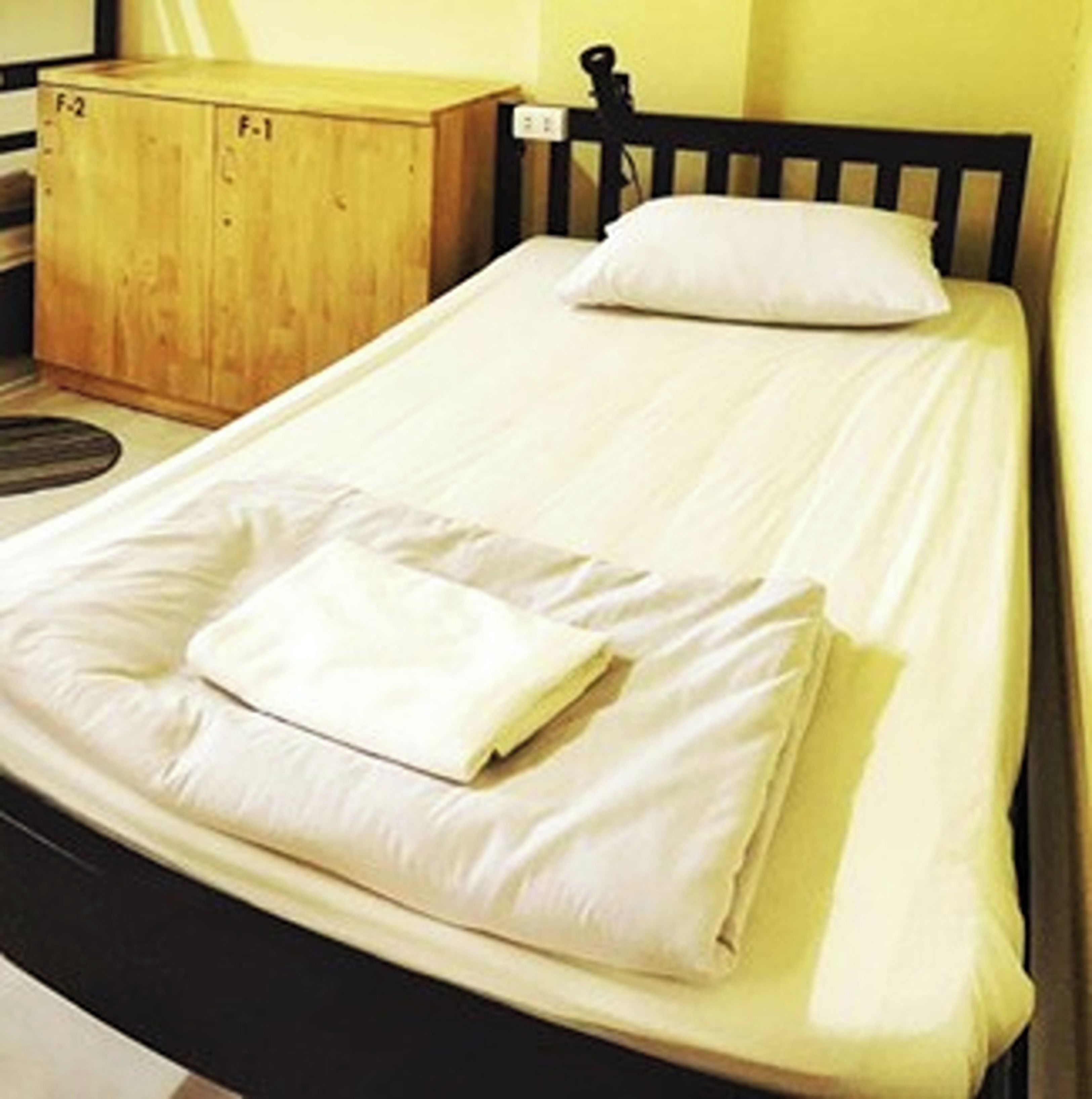 A single bed with white bedding and a folded white towel, a wooden nightstand with a lamp, and a black headboard.