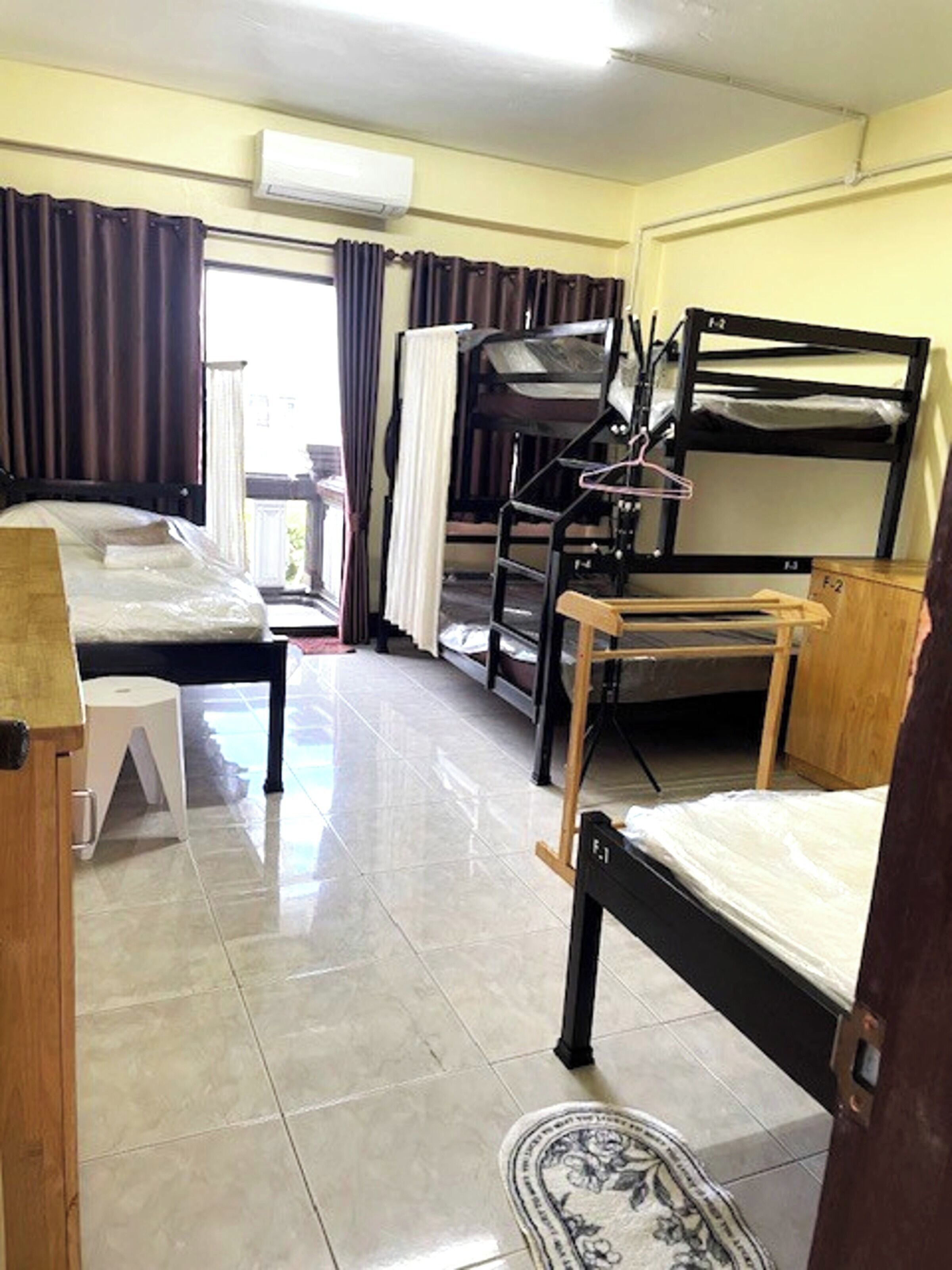 A dormitory room with bunk beds, a small table, a chair, a white stool, and a window with curtains.