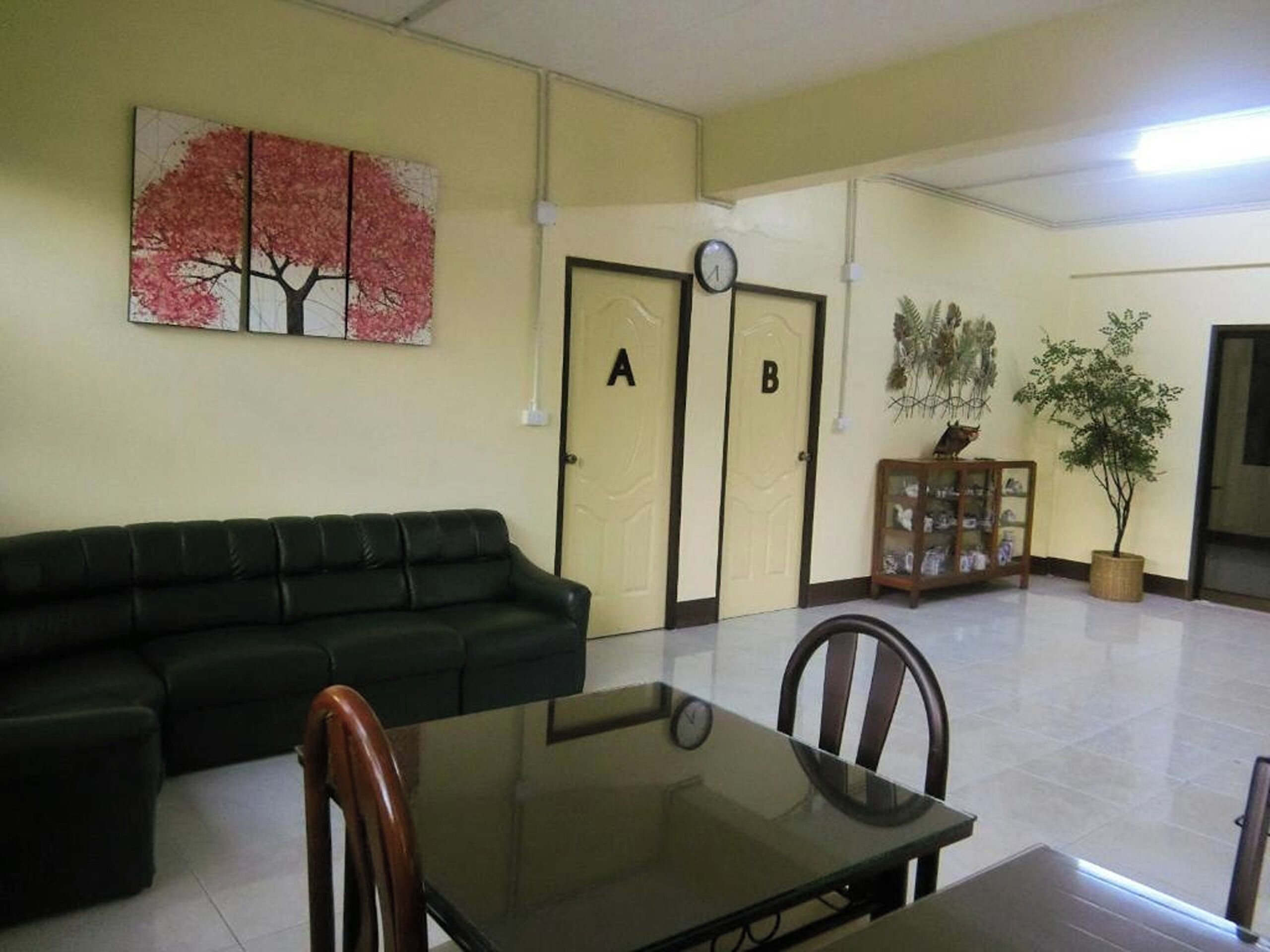 Una habitación con un sofá de cuero negro, una mesa de vidrio, sillas de madera, un reloj, una puerta con la etiqueta 'A', una puerta con la etiqueta 'B' y un cuadro colgado en la pared.