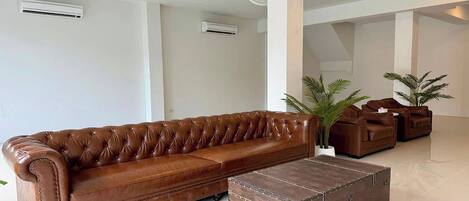 Un sofá de cuero marrón, un cofre de madera y dos plantas en macetas en una habitación moderna, bien iluminada, con paredes y ventiladores de techo blancos.