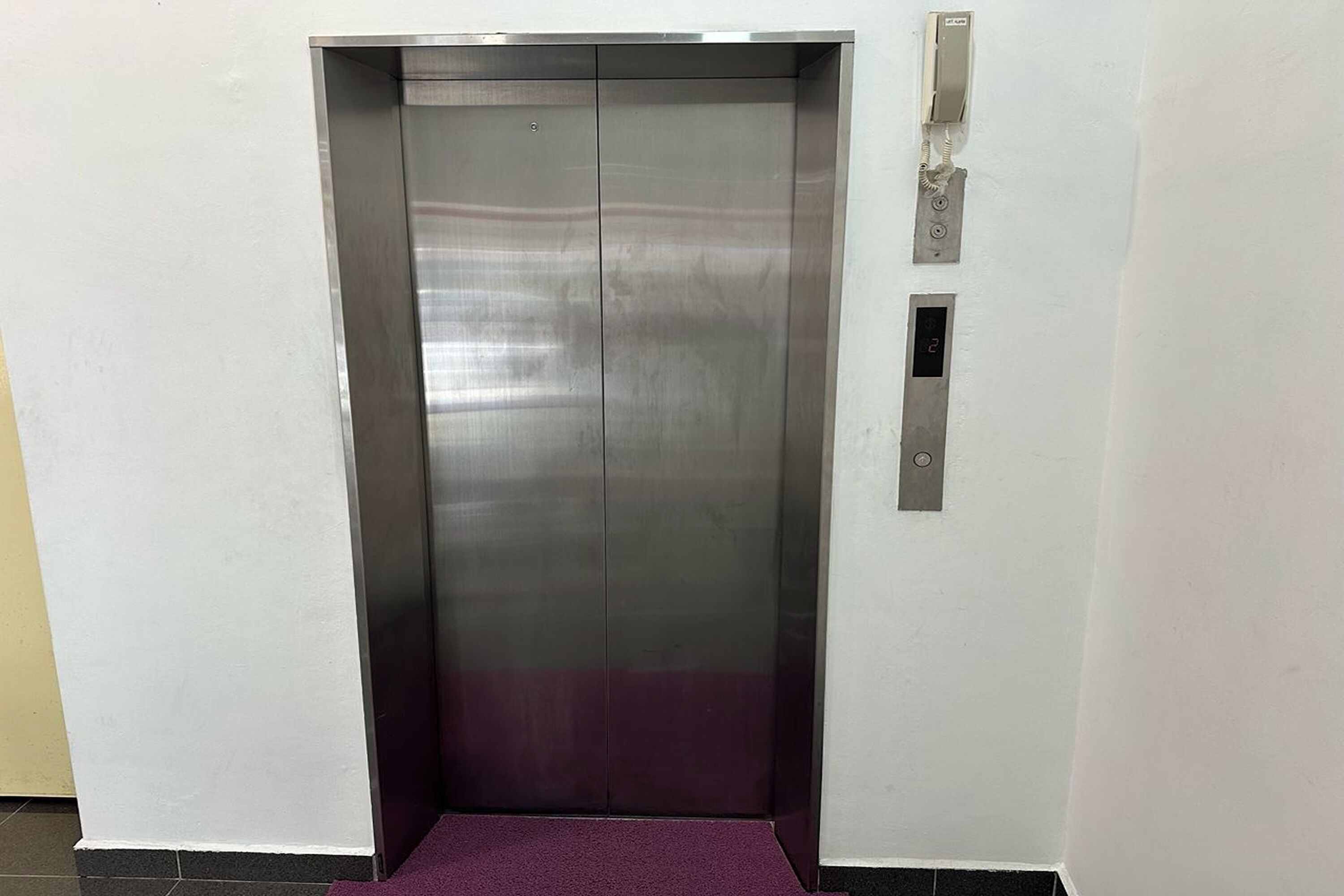 A metallic elevator with a purple carpet, a wall-mounted intercom, and a small ventilation panel.