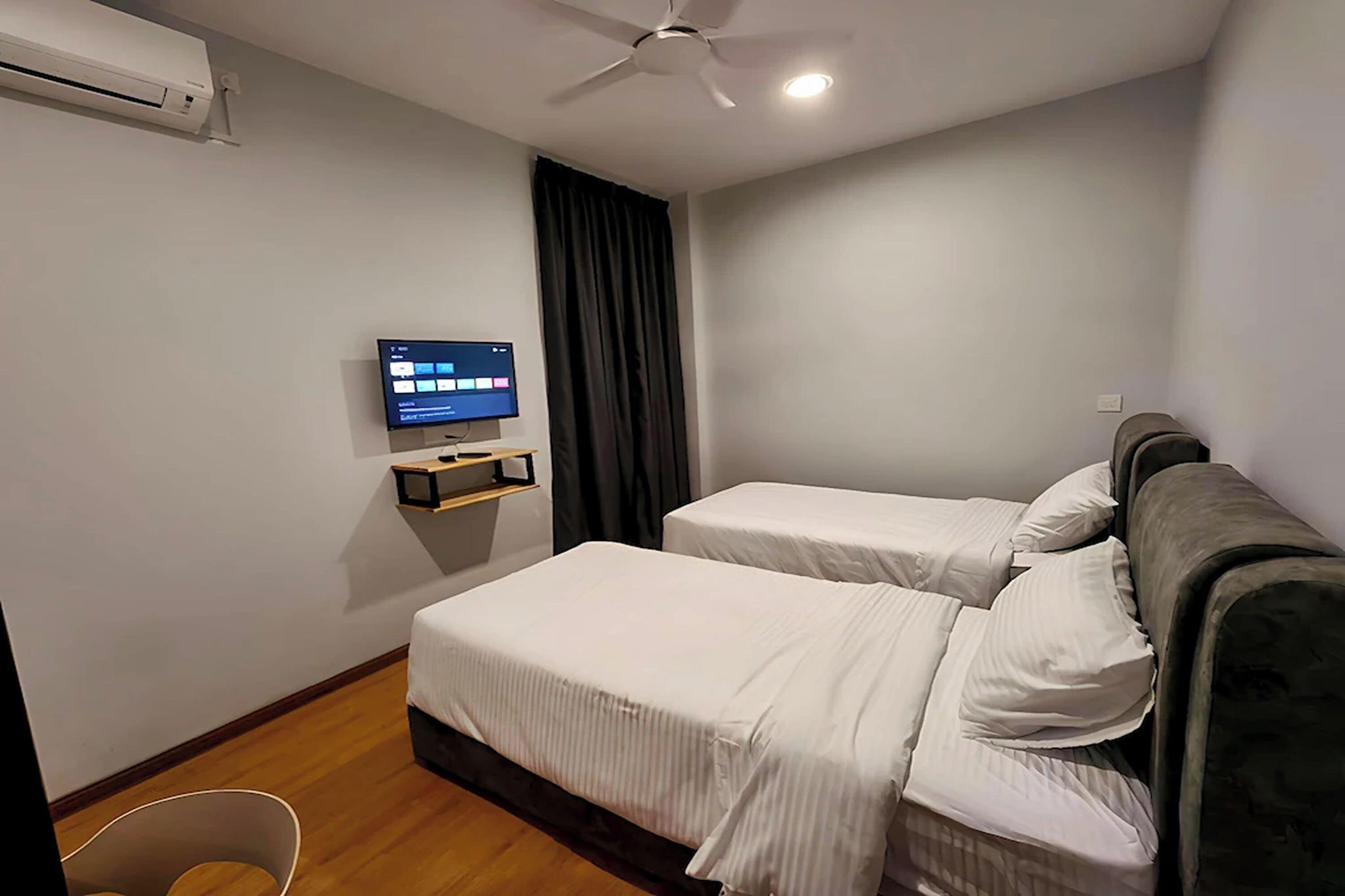 A hotel room with two beds, a ceiling fan, an air conditioning unit, a TV on a shelf, and a dark curtain.
