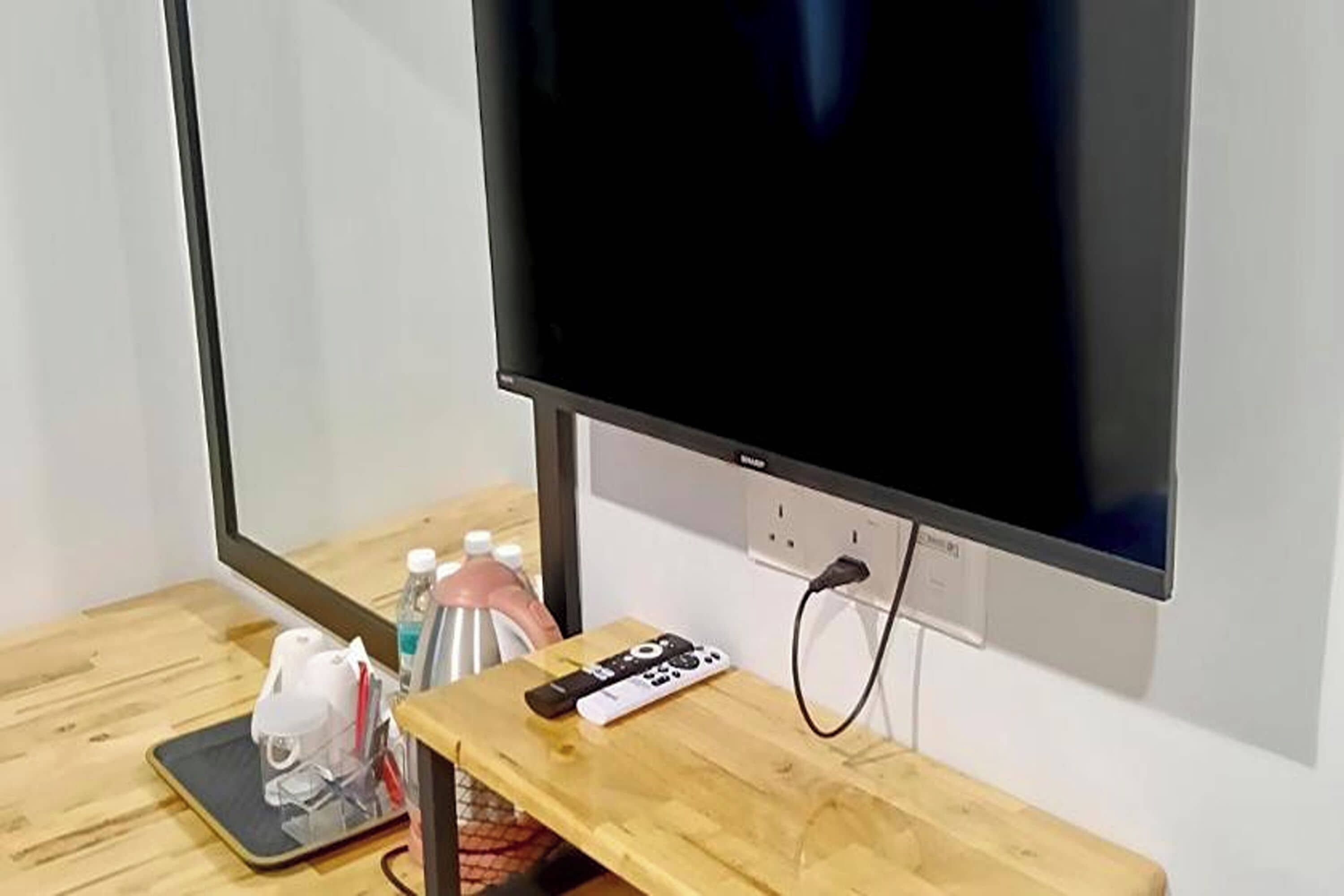 A TV mounted on a wall, a wooden table with a remote control, a power outlet, and a small shelf with bottles.