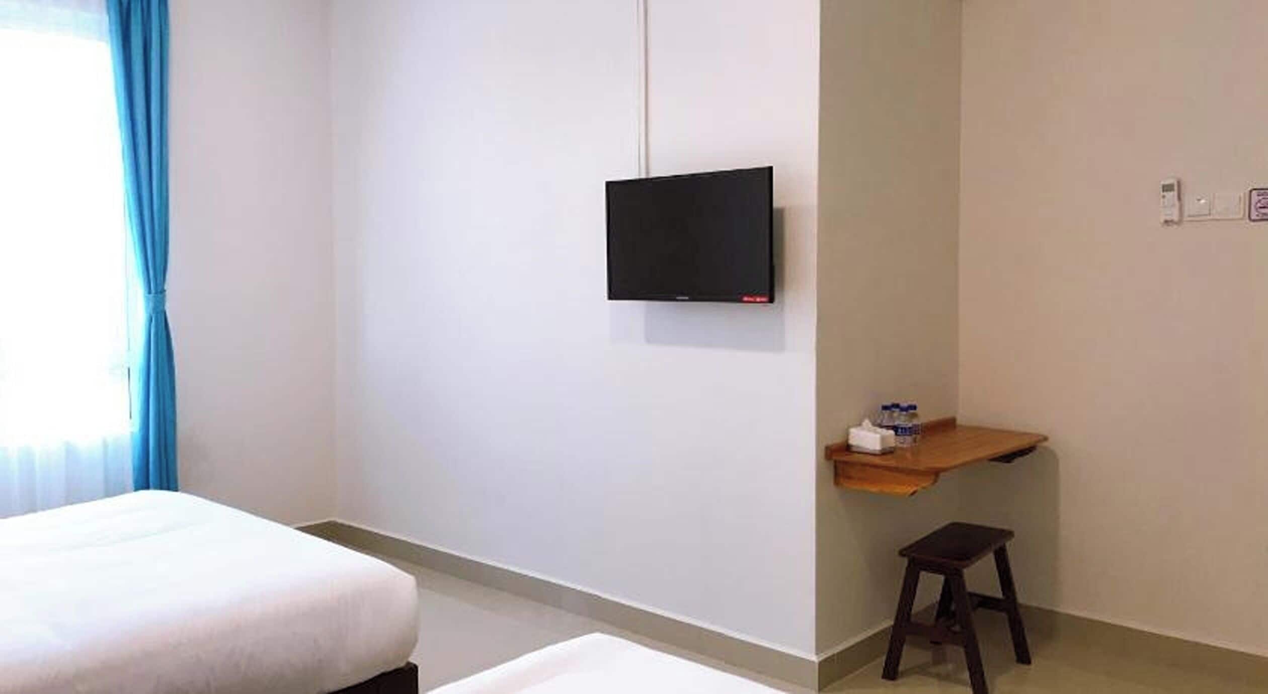 A hotel room with a bed, a desk with a chair, a television mounted on the wall, and a window with curtains.