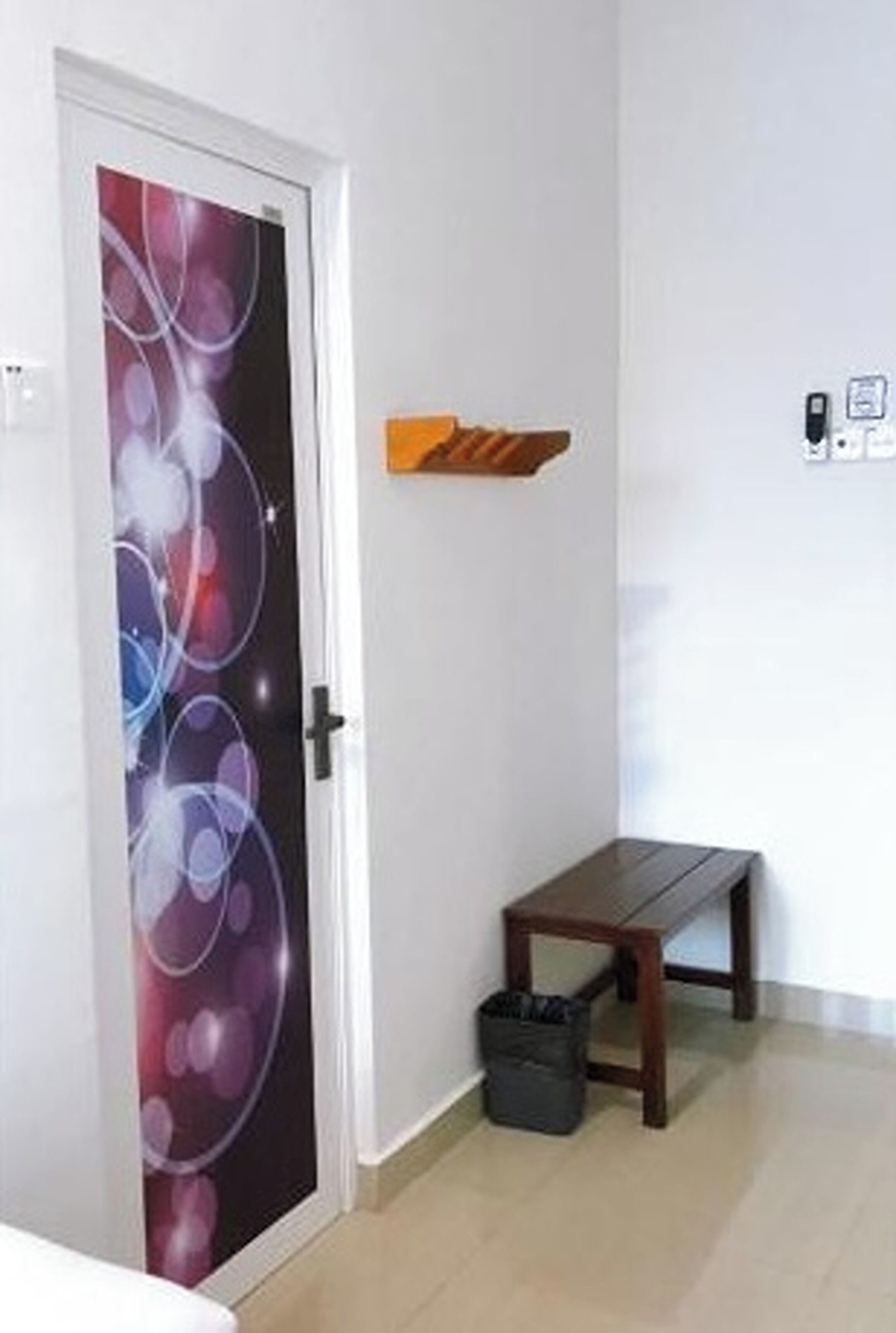 A room with a door featuring a colorful abstract design, a small wooden bench, and a wall-mounted shelf.