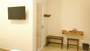 Um quarto pequeno com televisão fixada na parede, um banco, uma mesinha com garrafas e uma lixeira.