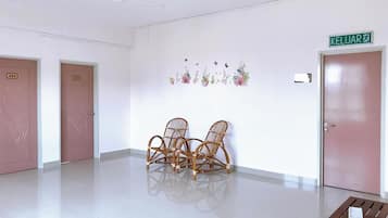 Een gang met twee deuren, waarvan er één '403' aangeeft en de ander een groen bordje met de tekst 'KELUAR' heeft. In het midden staan rieten stoelen en aan het einde van de gang een kleine tafel.