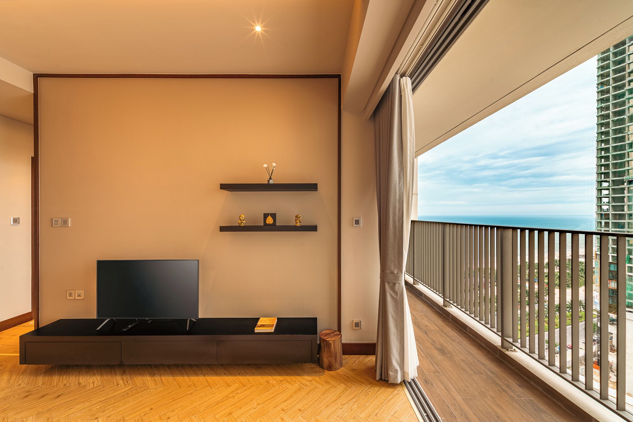 A modern living room with a flat-screen TV, wooden flooring, and a balcony with a view of the city.