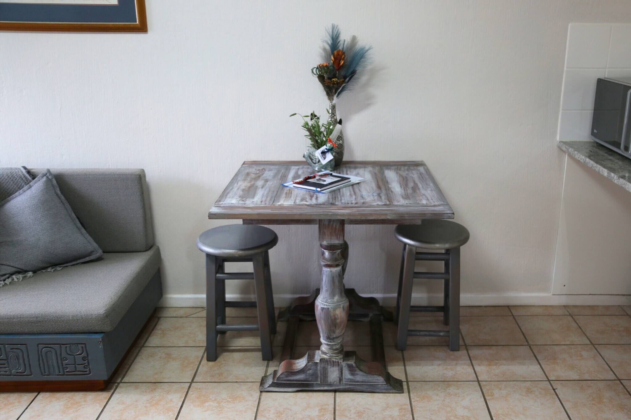 A small dining area with a wooden table, two stools, a vase with flowers, and a gray sofa with cushions.