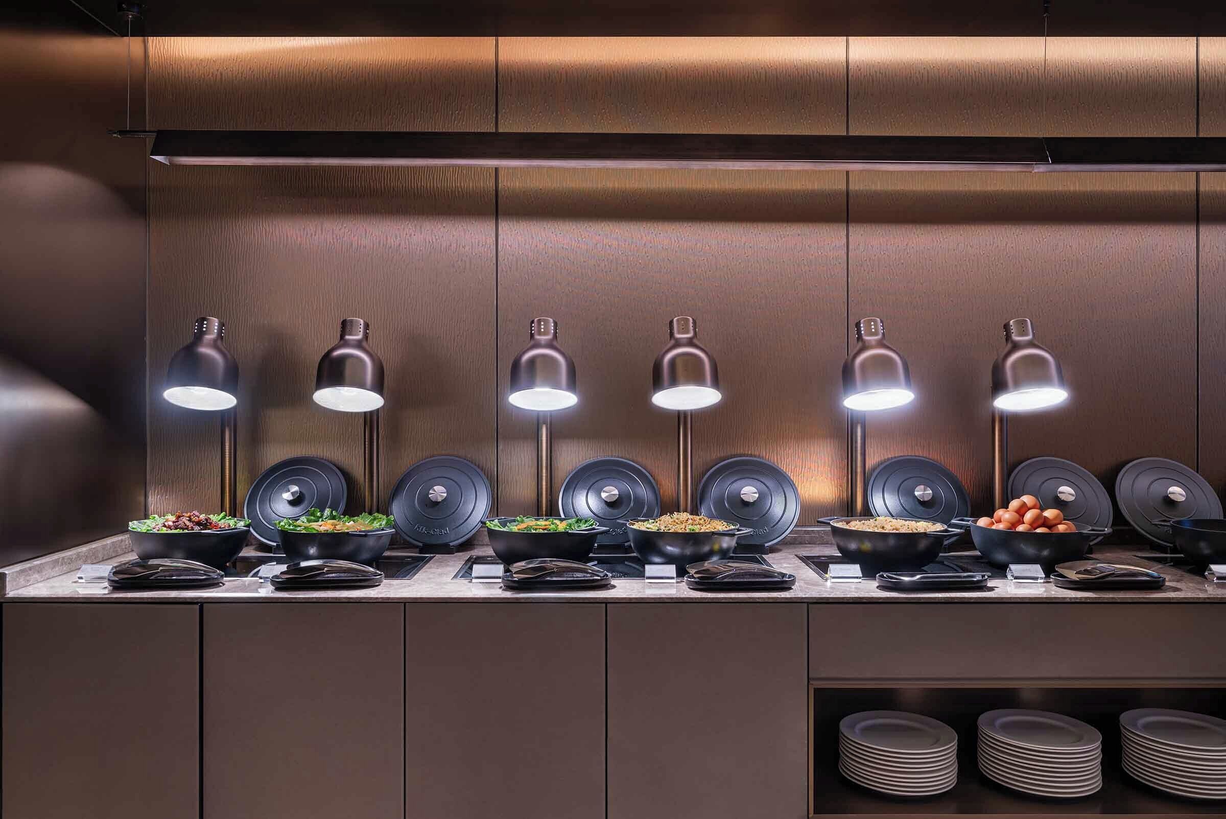A buffet setup with multiple food stations, overhead lighting, and stacked plates.