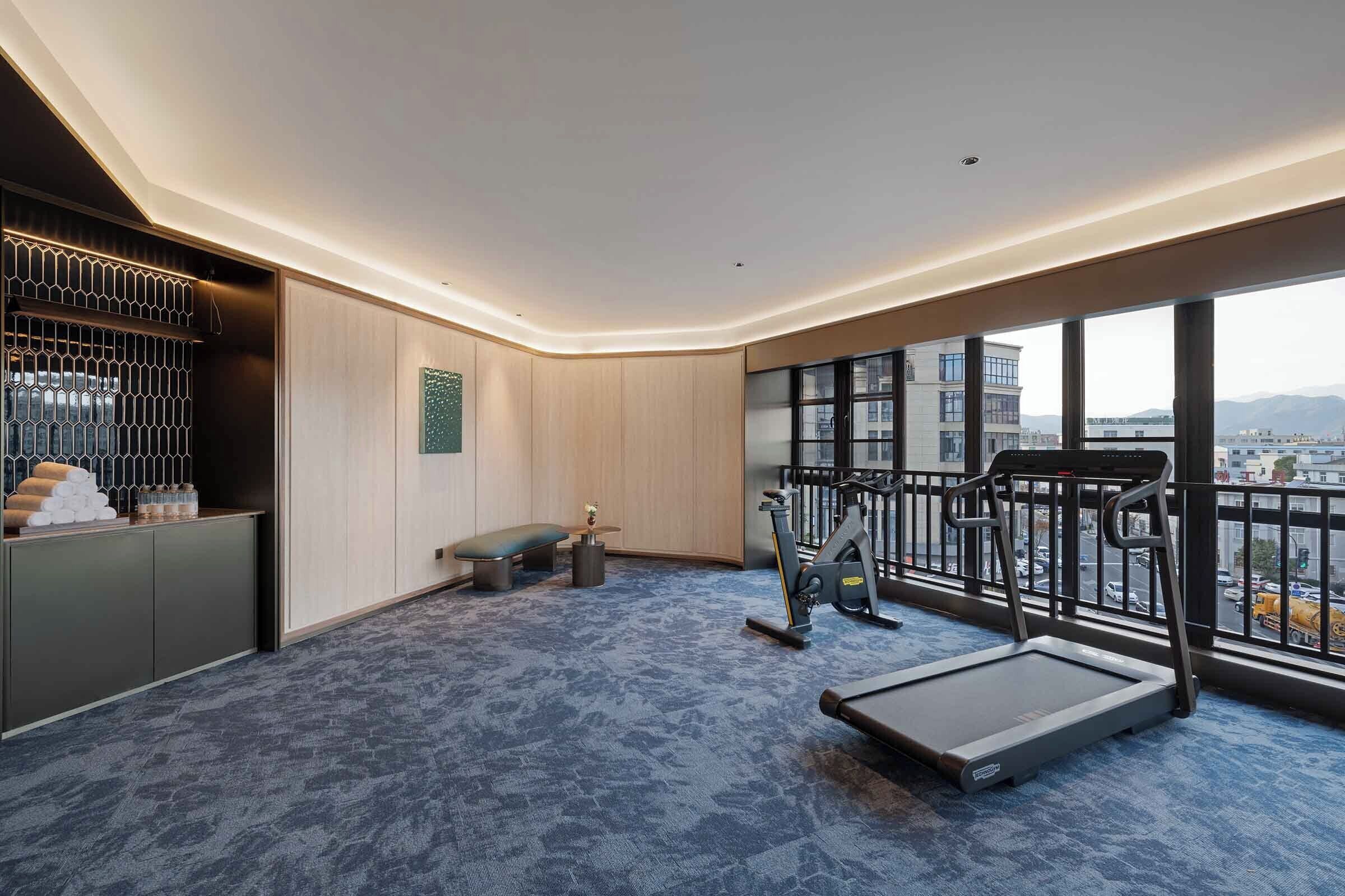 A hotel room with a large window, a treadmill, a bench, and a small exercise area.