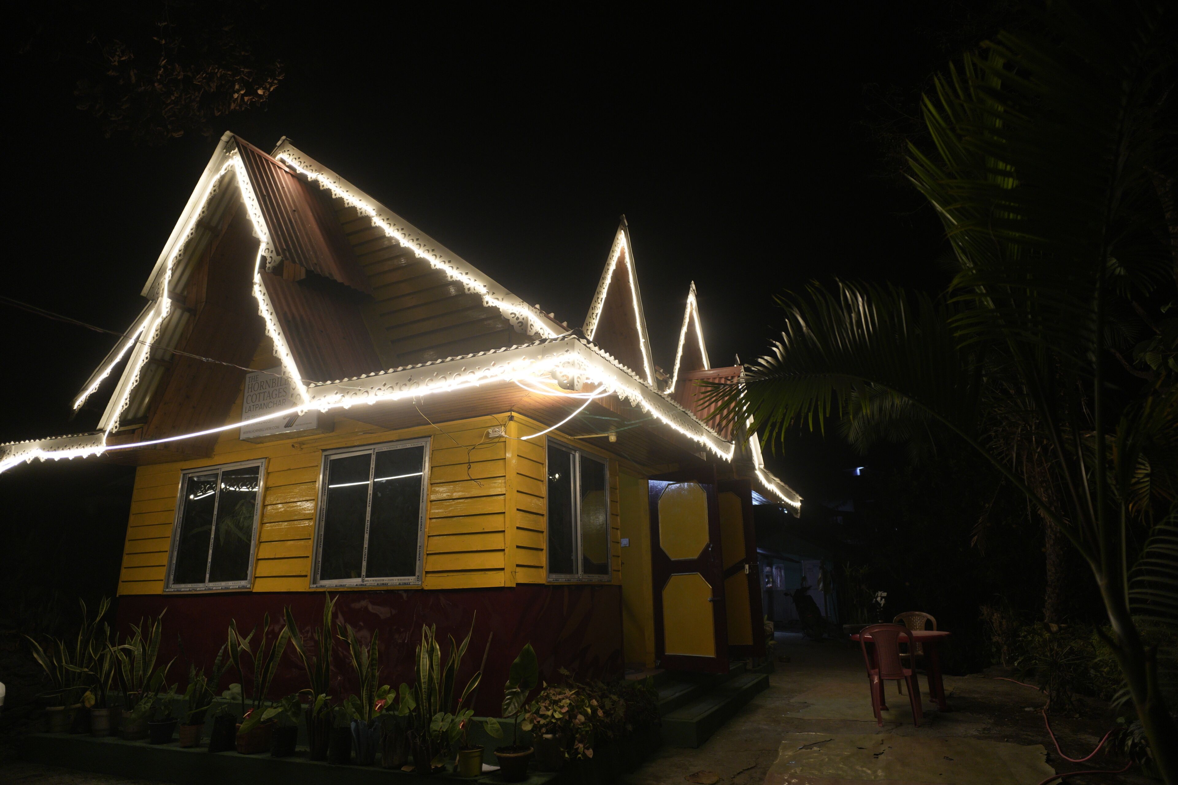 A yellow building with a sloped roof, surrounded by potted plants.