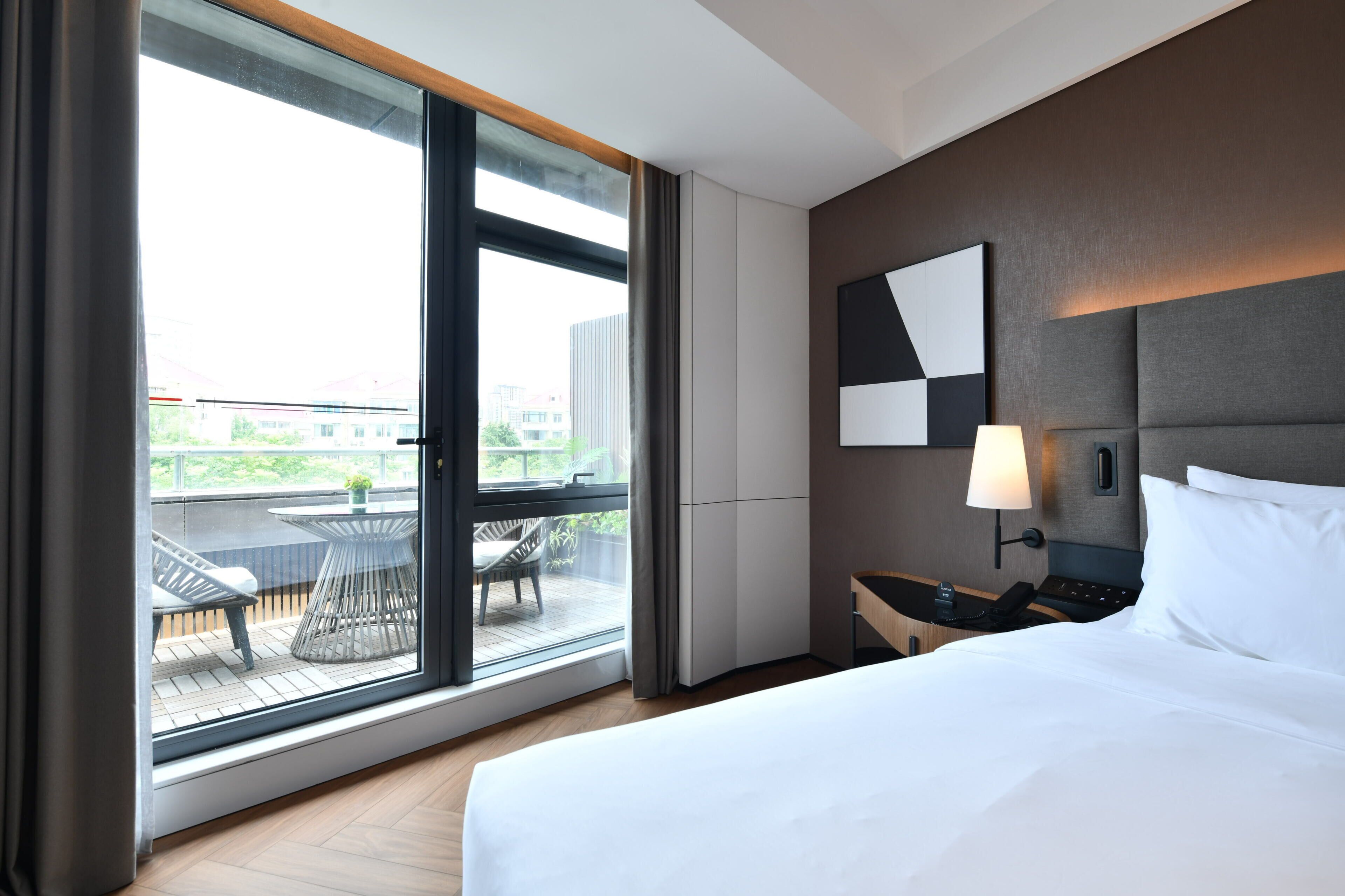 A hotel room with a large window, a bed, a nightstand, a lamp, and a view of the cityscape.