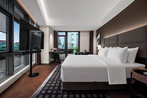 A modern hotel room with a large bed, a desk, a chair, a TV, and a view of the city.