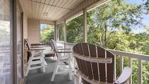 A covered porch with wooden chairs and tables, large windows, and a view of trees. - True Blue Golf & Beach Getaway (Pawleys Island)