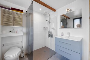 A modern bathroom with a glass shower enclosure, a white toilet, a vanity with a mirror, and a shelf with toiletries. - Charming Apartment for 4 with Shared Terrace and Garden (Saint-Jean-de-Liversay)