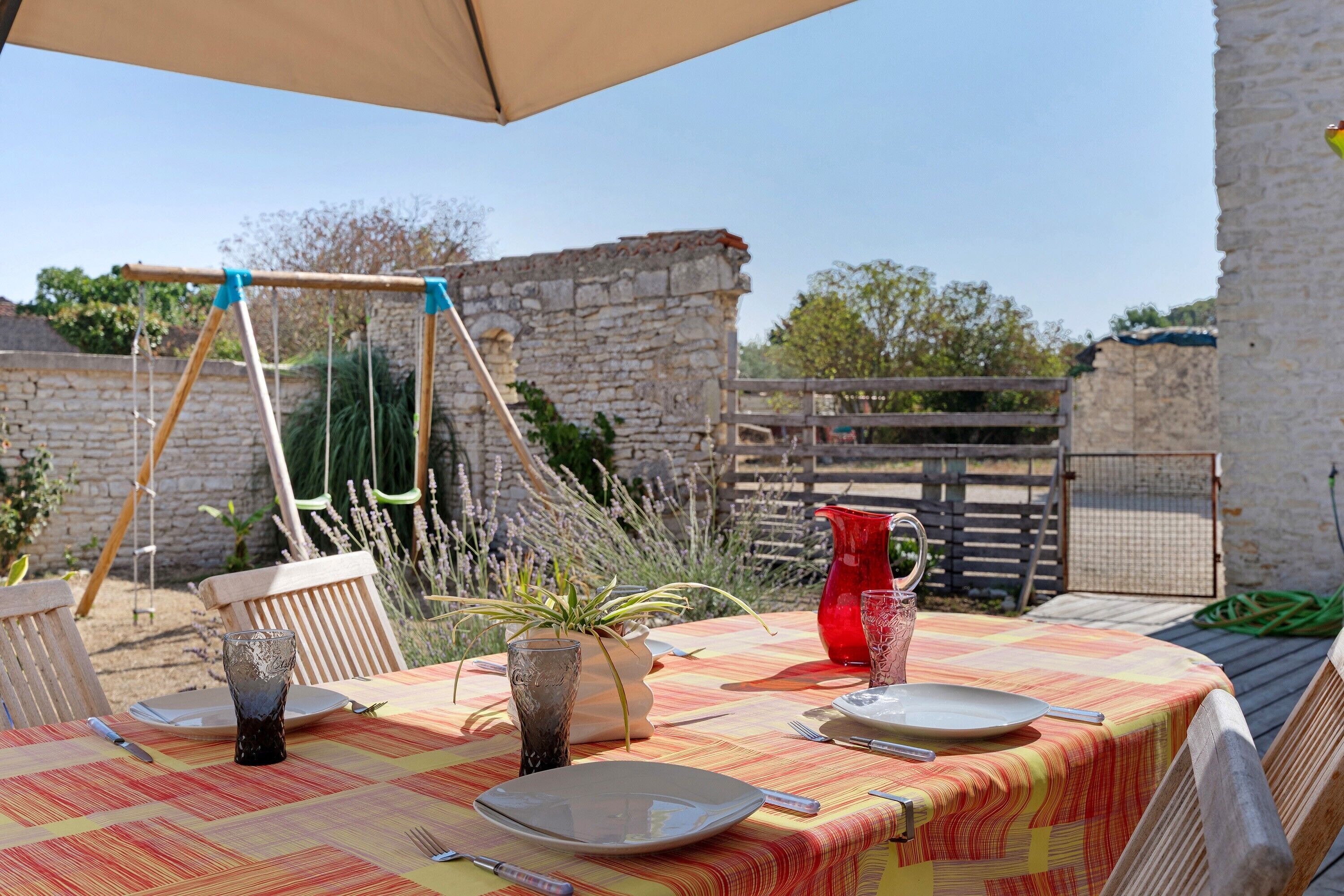 Un espace extérieur aménagé pour les repas, avec une table dressée pour deux personnes, une balançoire et un mur de pierre en arrière-plan.