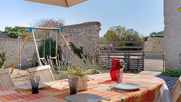 Un espace extérieur aménagé pour les repas, avec une table dressée pour deux personnes, une balançoire et un mur de pierre en arrière-plan.