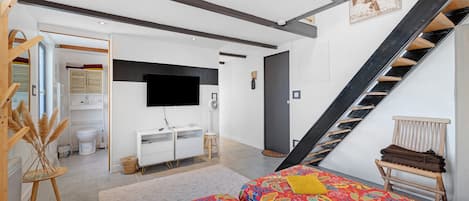 Un dormitorio moderno con cama, televisor, escalera y un baño visible al fondo.