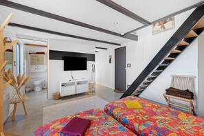 Un dormitorio moderno con cama, televisor, escalera y un baño visible al fondo.