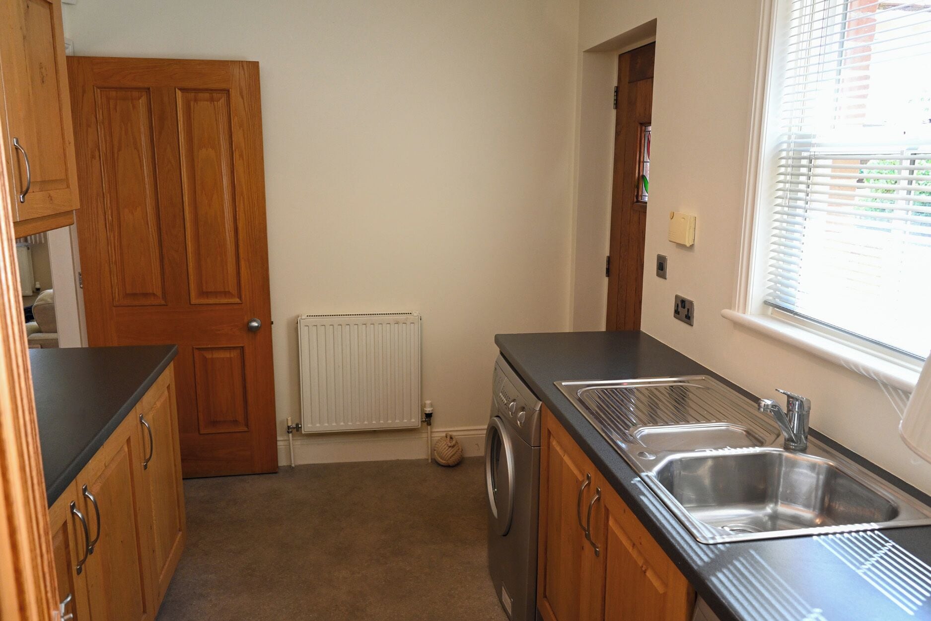 Une cuisine avec des meubles de rangement en bois, un lave-linge, un radiateur et une fenêtre avec des stores.