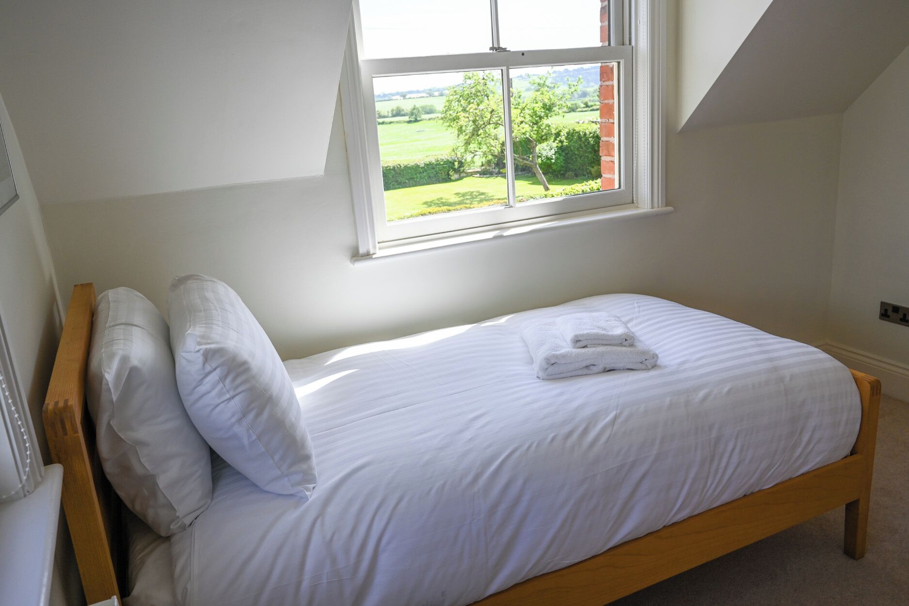 A single bed with white bedding, a window with a view of greenery, and a wooden headboard.