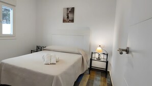 Een eenpersoonsbed met een witte spreideken en twee opgerolde handdoeken, een nachtkastje met een lamp, een raam met uitzicht op bomen, en een schilderij aan de muur.