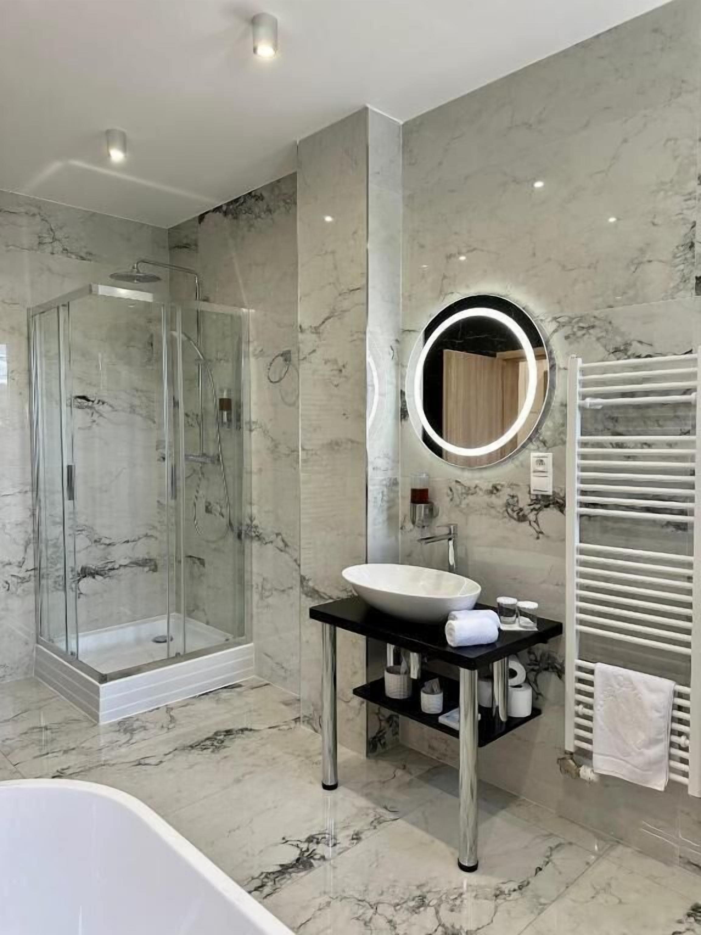 A modern bathroom with a glass shower enclosure, a round mirror, a white sink, and a towel rack.