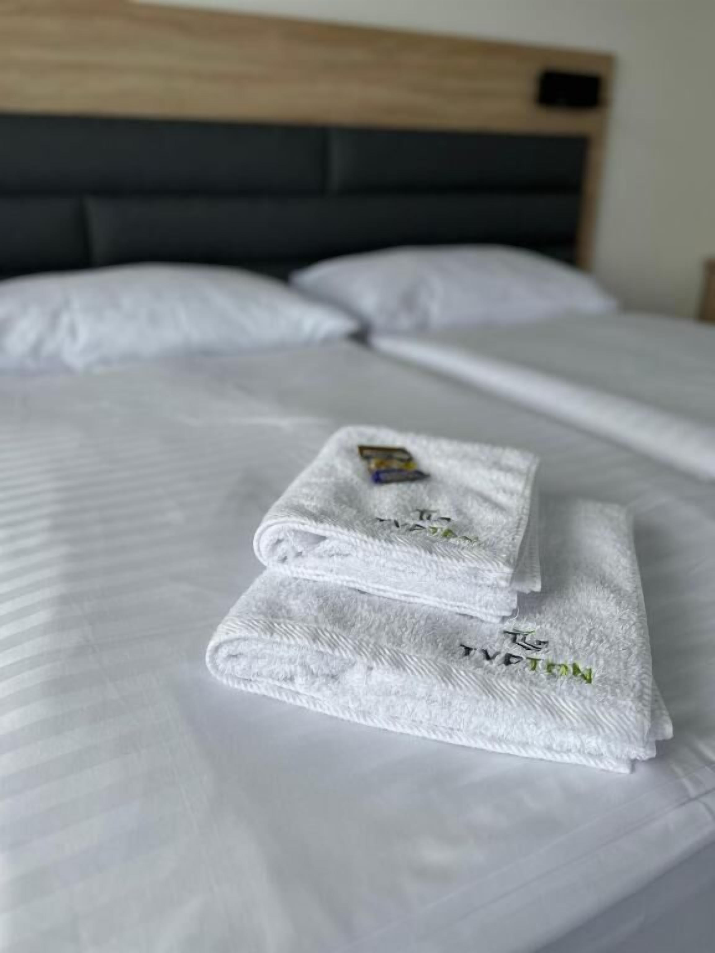 A neatly made bed with white linens and two folded towels with embroidered text.