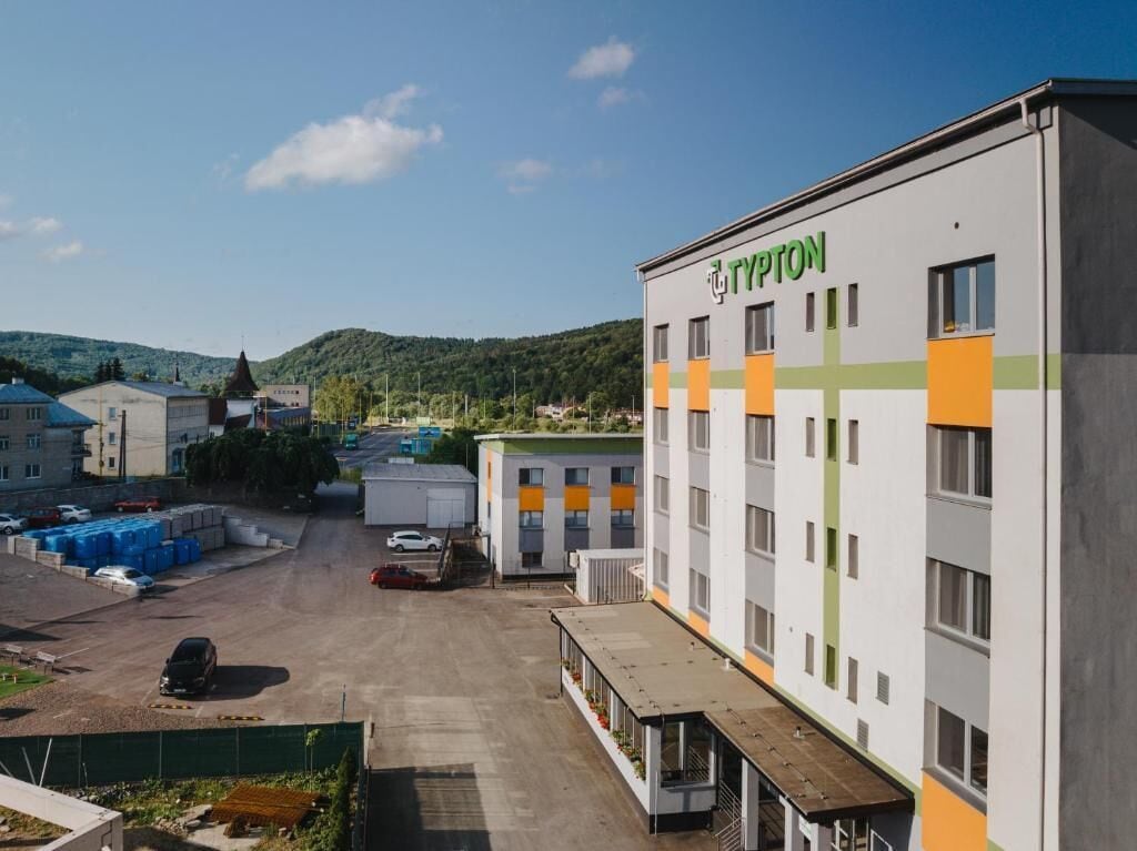 A Typton hotel with a parking lot, a green hill in the background, and a clear sky.