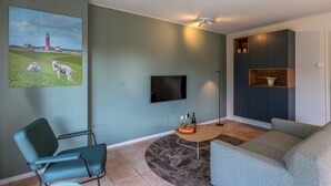 Una sala de estar con una televisión de pantalla plana, un sofá, una silla, una mesa de centro y un cuadro de un faro y ovejas.