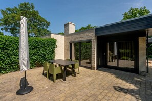 Eine Terrasse mit Tisch und Stühlen, einem schattenspendenden Sonnenschirm und einem modernen Gebäude mit großen Fenstern und einer Ziegel-Fassade.