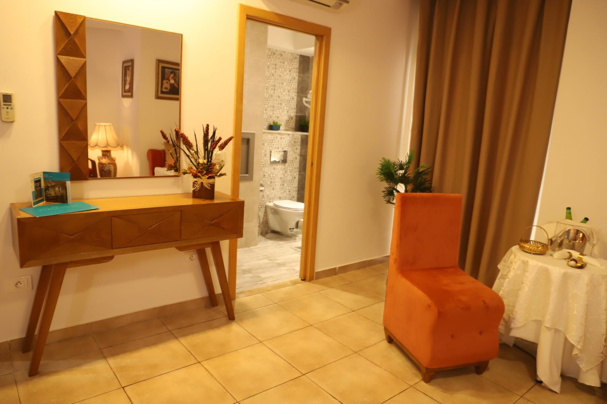 A hotel room with a wooden console desk, a small table with a white tablecloth, an orange armchair, and a bathroom visible through an open door.