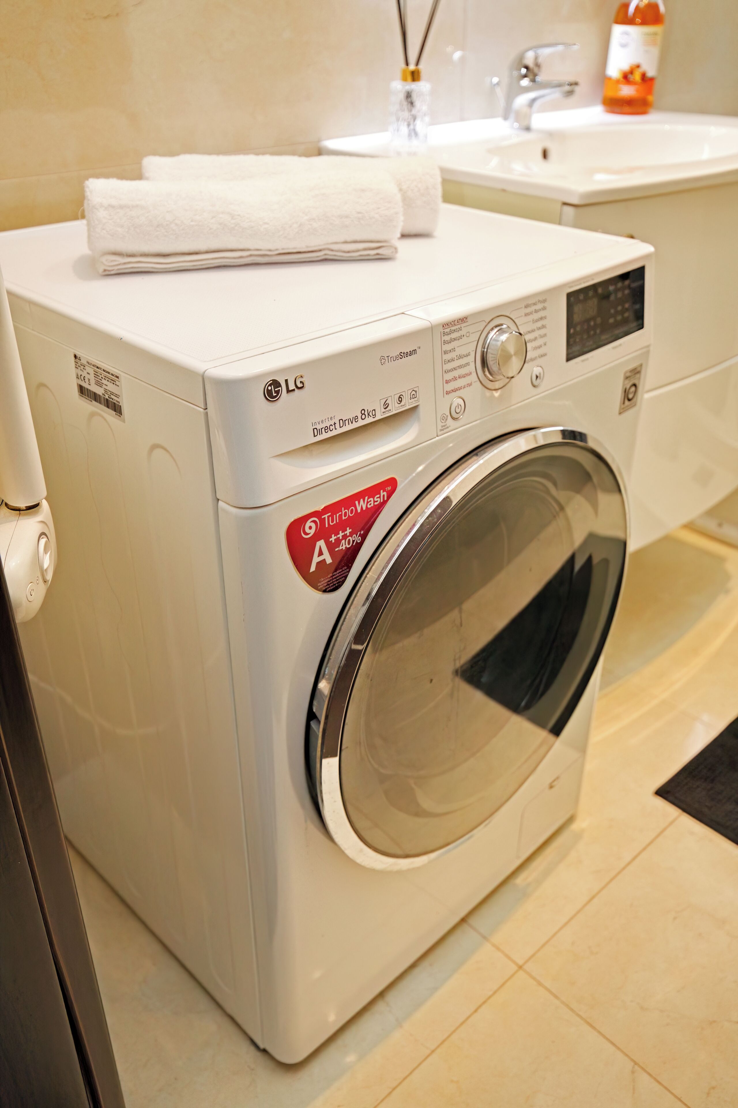 Une machine à laver LG à entraînement direct blanche, modèle 85, dotée de la technologie TurboWash A+40x, placée dans une salle de bain avec un lavabo et un flacon de savon liquide pour les mains.